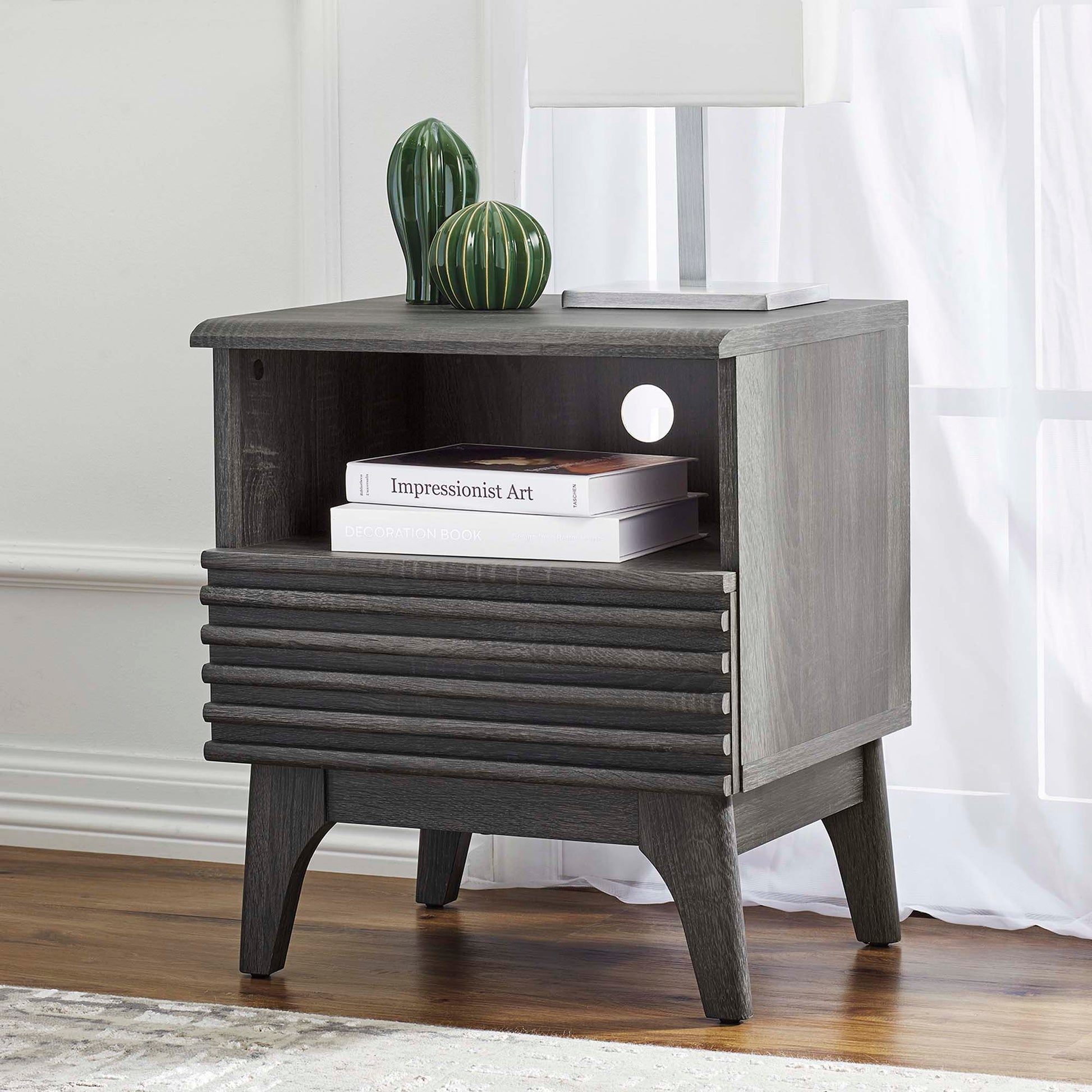 Black nightstand with a slatted drawer front, open shelf, and tapered legs; green ceramic vases and a lamp on top.