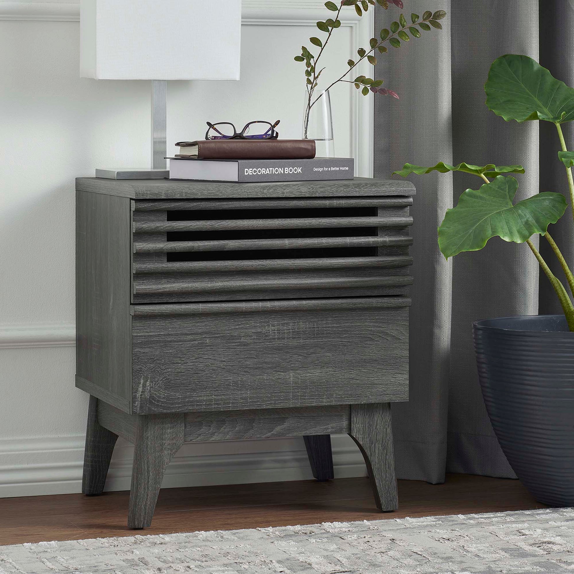 Black mid-century wood nightstand with a slatted front, shown with a lamp, books, and a tall plant in a bedroom