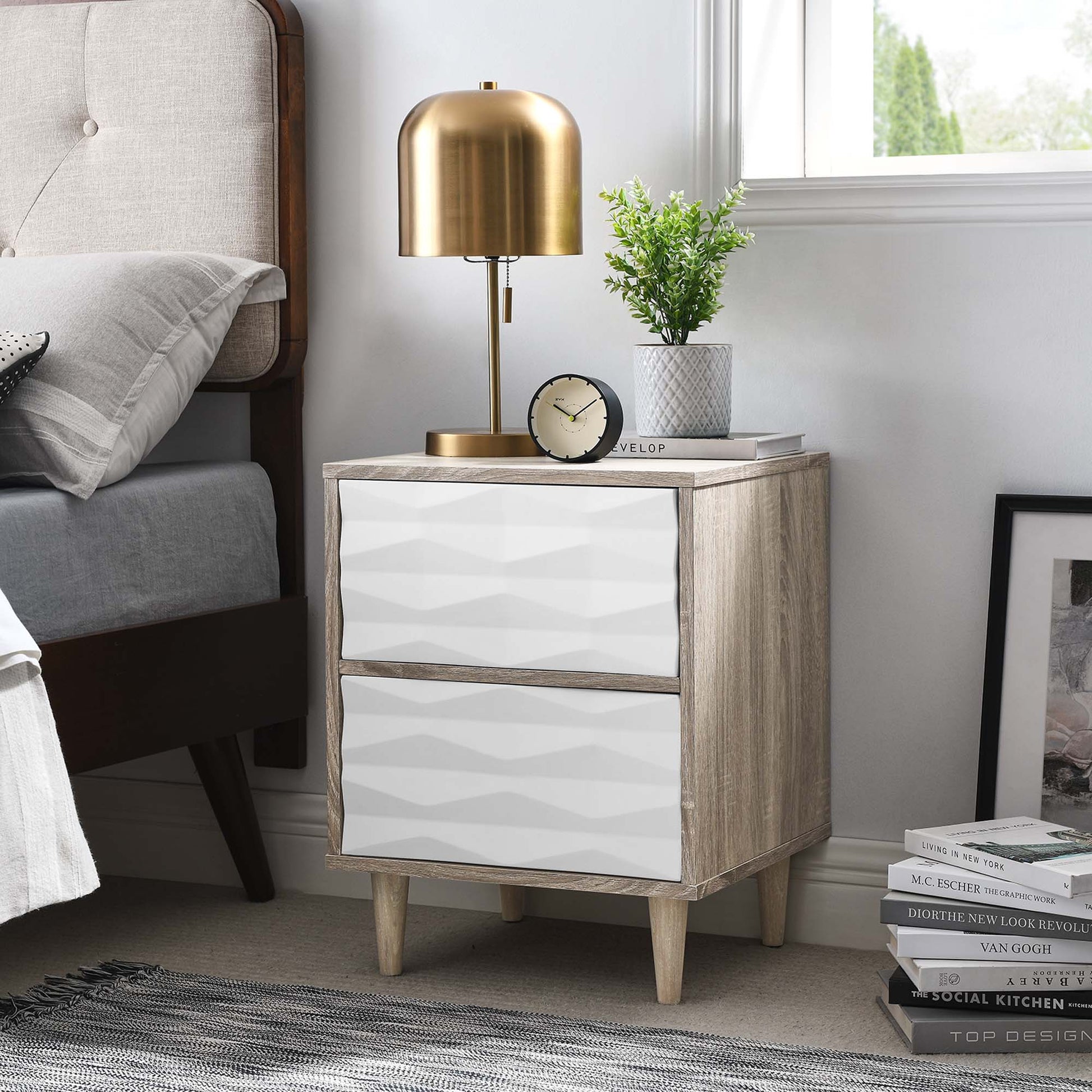 Oak nightstand with two white textured drawer fronts and tapered legs, plus brass lamp, clock, plant, and books on top.