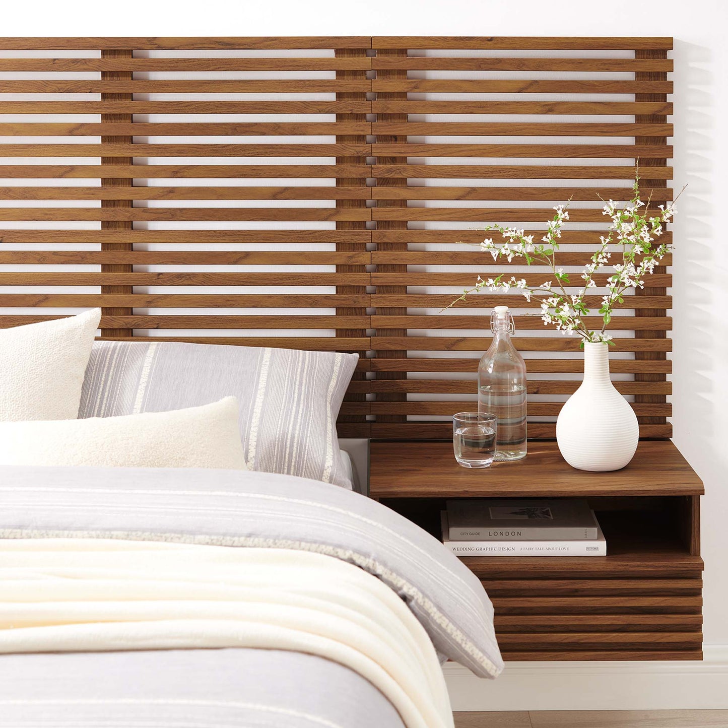 Walnut slatted wall-mounted twin headboard with floating nightstands, glass bottle, white vase with blossoms, cozy gray-cream bedding.