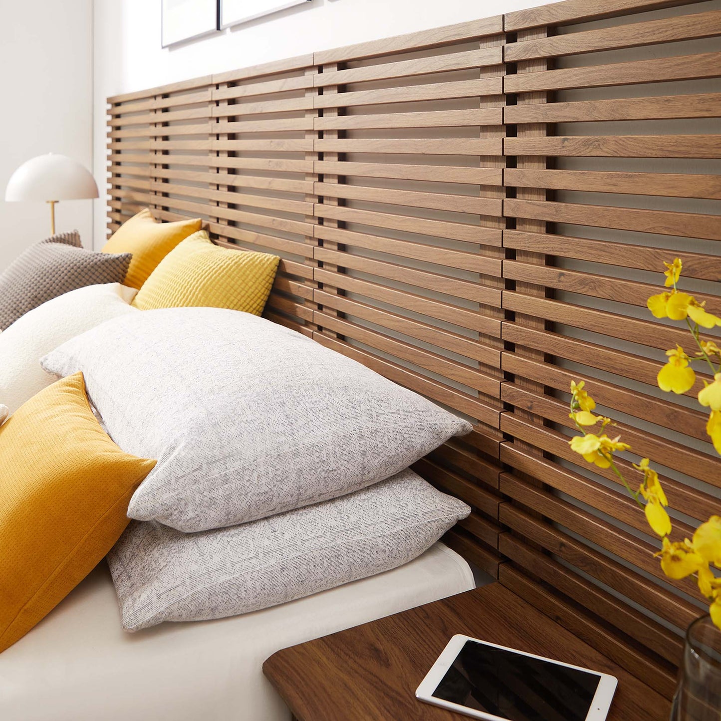 Walnut slat wall-mounted queen headboard with modern nightstands, bed with white bedding, gray and mustard cushions, bedside lamp and yellow flower vase.
