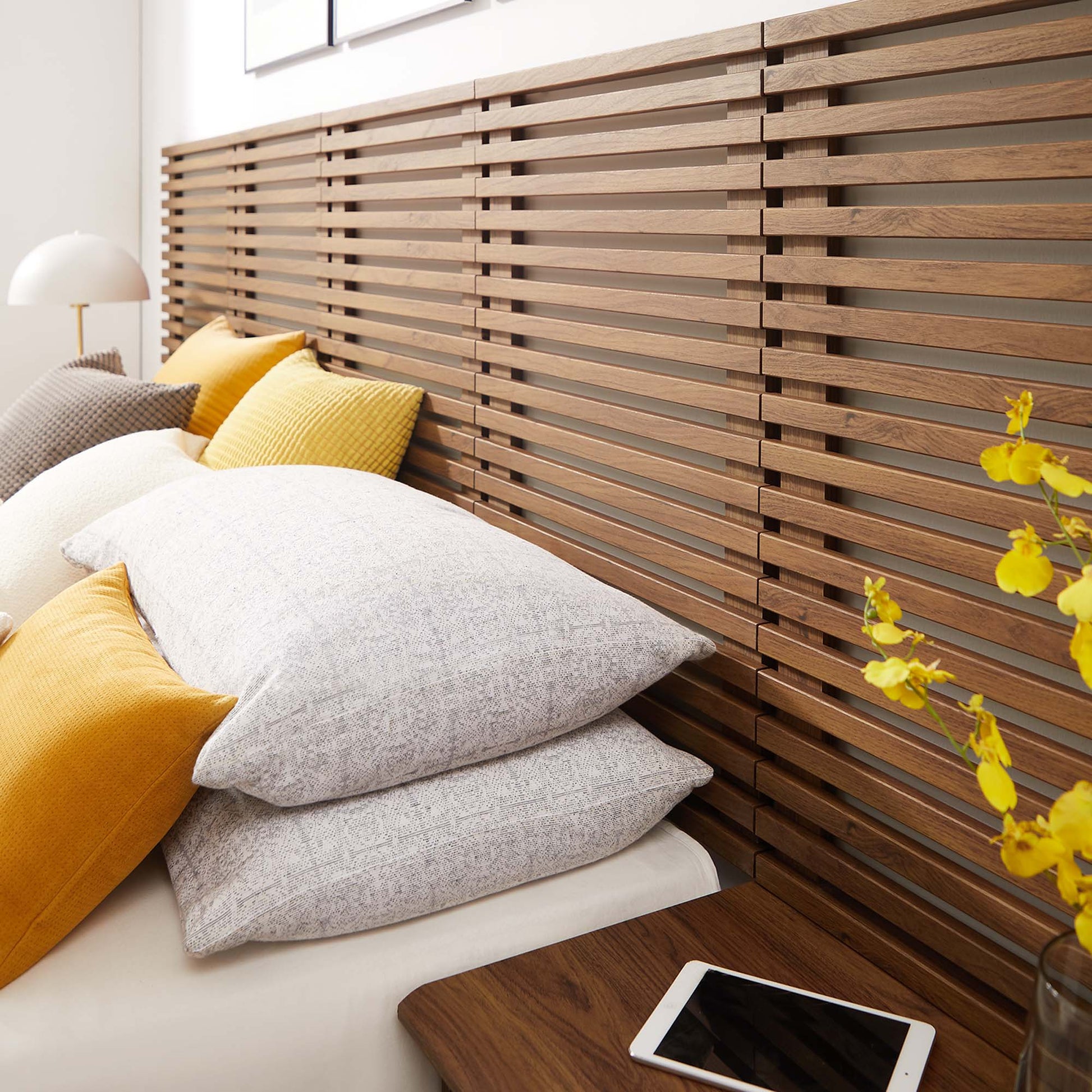 Walnut slat wall-mounted queen headboard with modern nightstands, bed with white bedding, gray and mustard cushions, bedside lamp and yellow flower vase.
