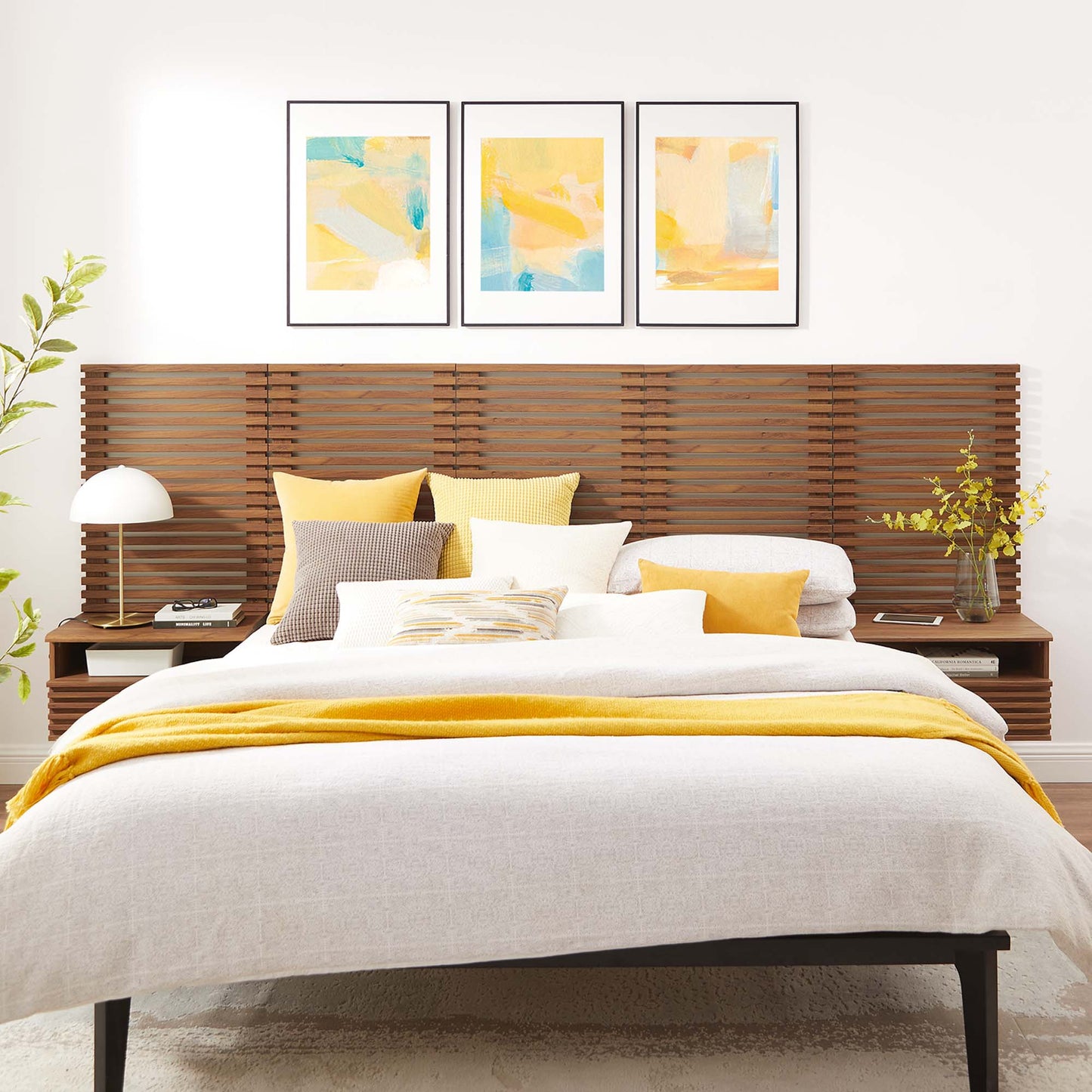 Walnut wall-mounted queen headboard with beige bedding, yellow throw and pillows; three pastel abstract prints above.