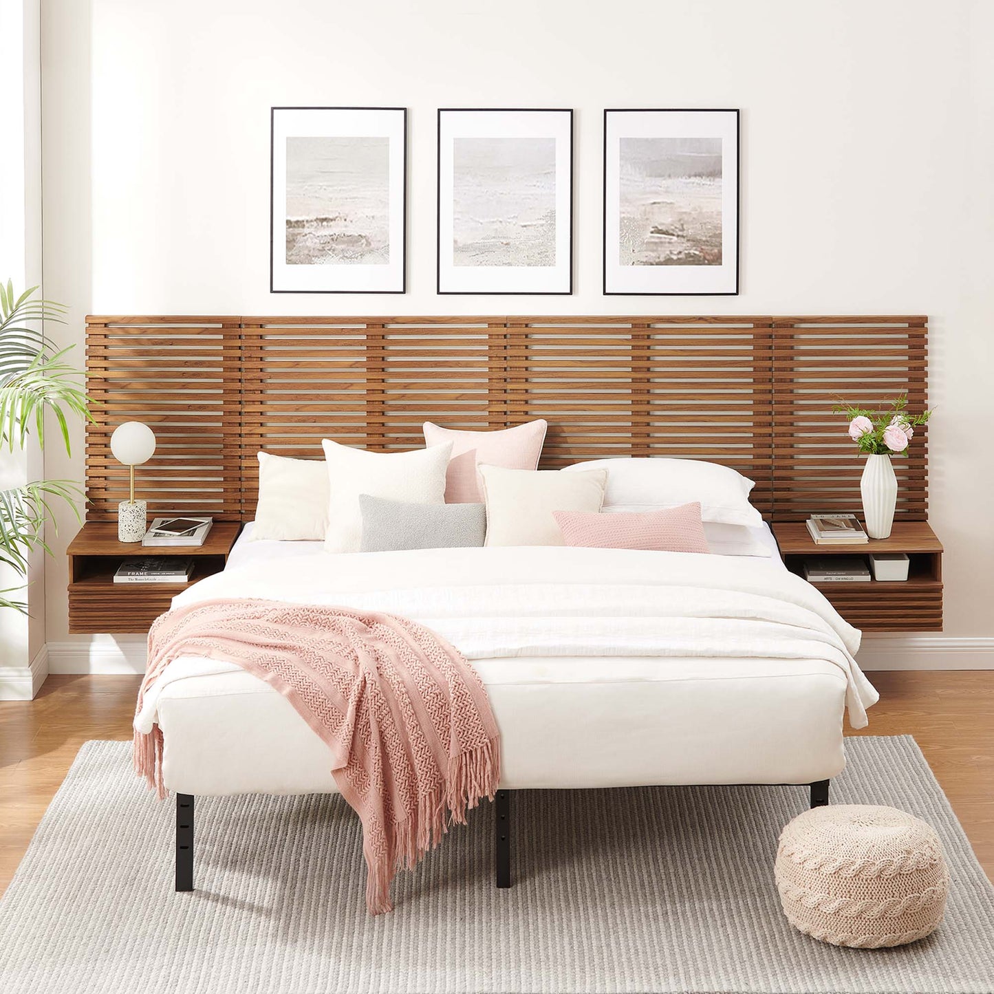 Walnut slatted, wall-mounted king headboard with modern nightstands; white bedding, pastel cushions, blush throw; seaside art, rug, knit pouf.