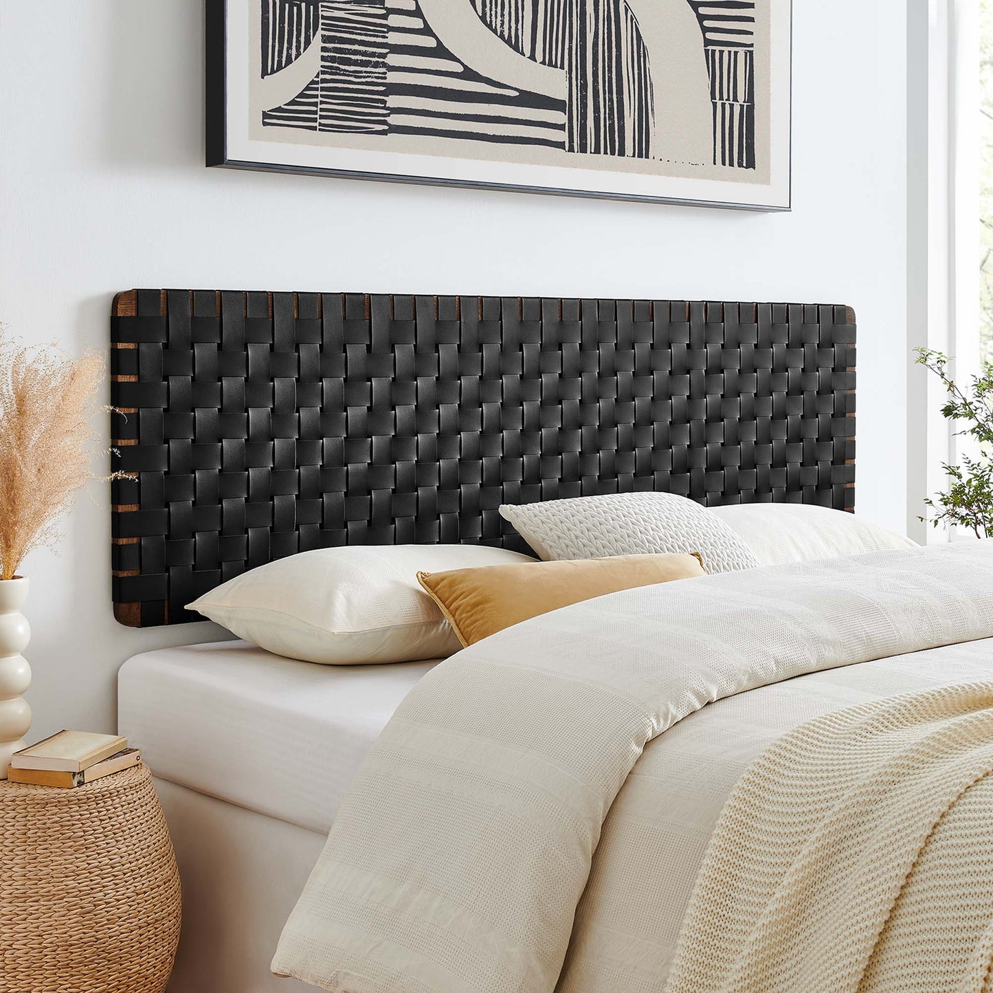Woven black wall-mount headboard (vegan leather) for a king bed; white bed with cream duvet and tan accent pillow; wicker nightstand and abstract art above.