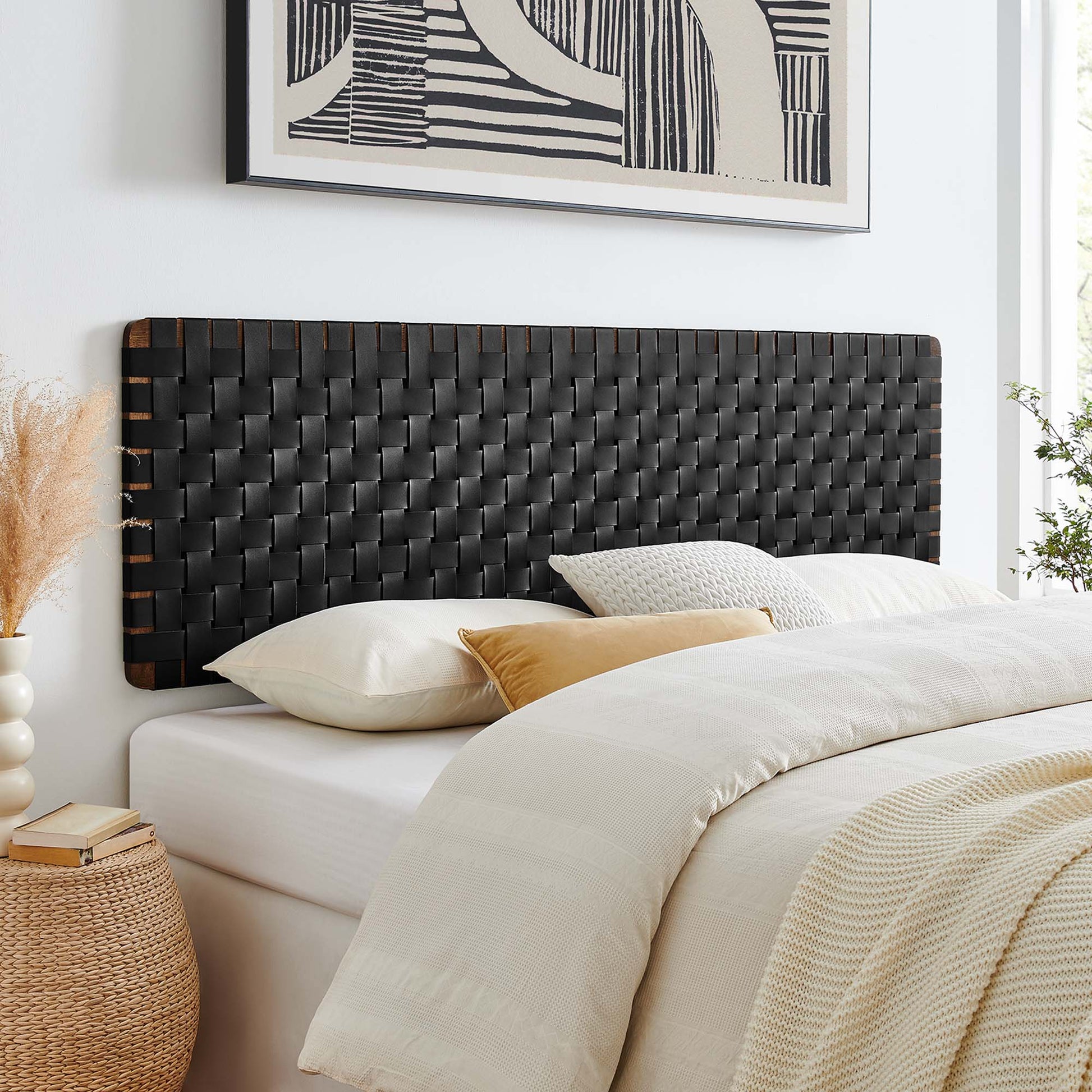 Woven black wall-mount headboard (vegan leather) for a king bed; white bed with cream duvet and tan accent pillow; wicker nightstand and abstract art above.