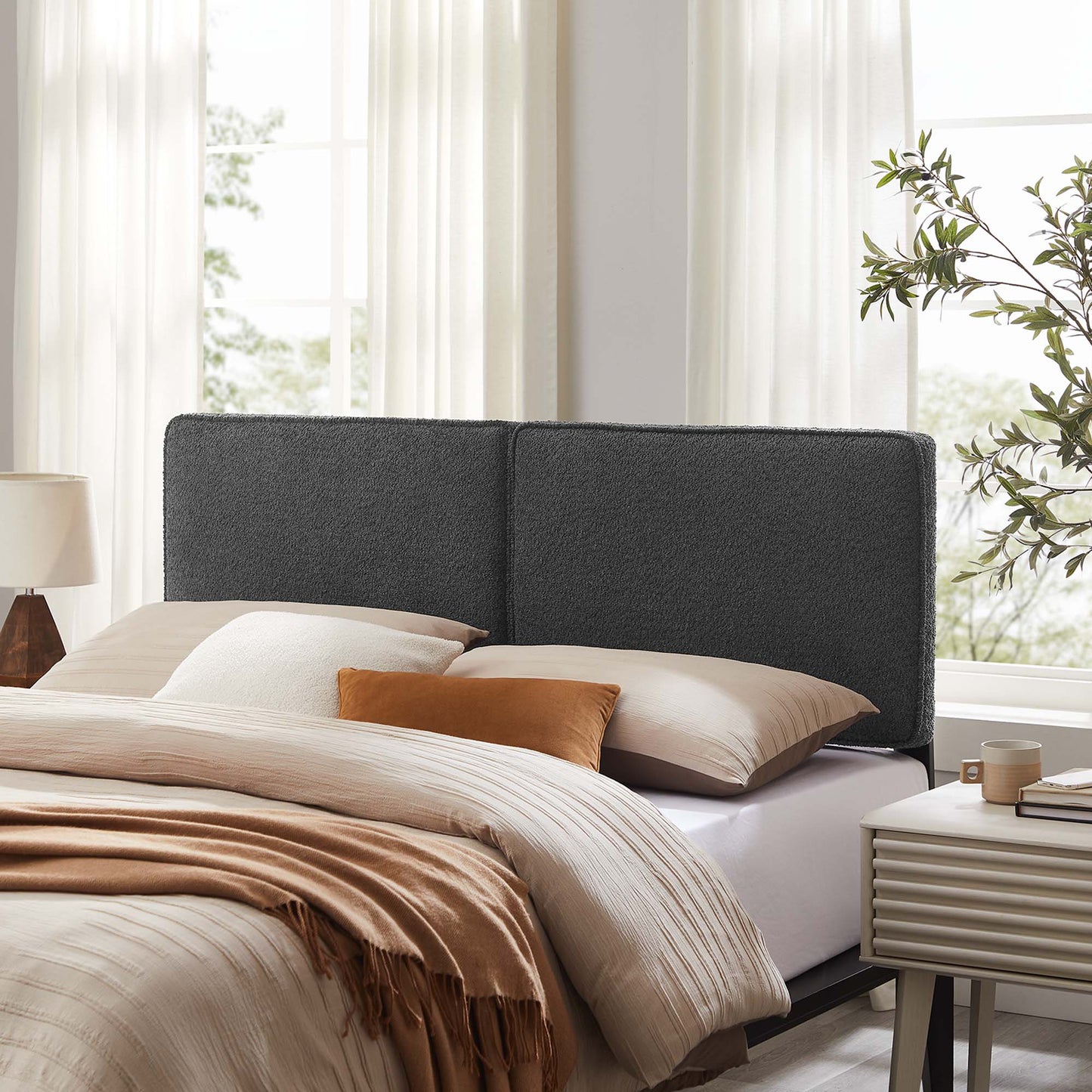 Dark gray upholstered bed with high headboard, beige sheets and tan throw, bright bedroom with nightstand and plant.
