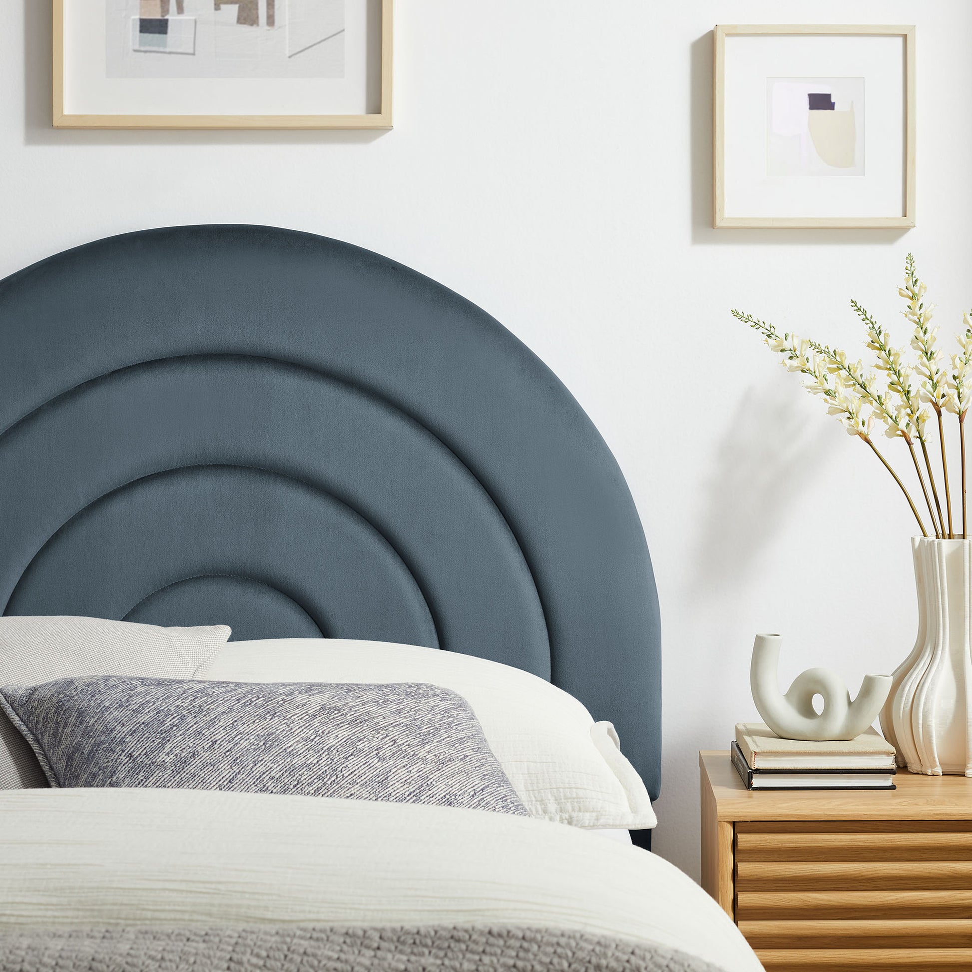 Circular navy upholstered headboard with concentric arches; white bedding, gray throw, wooden nightstand with vase of dried flowers.
