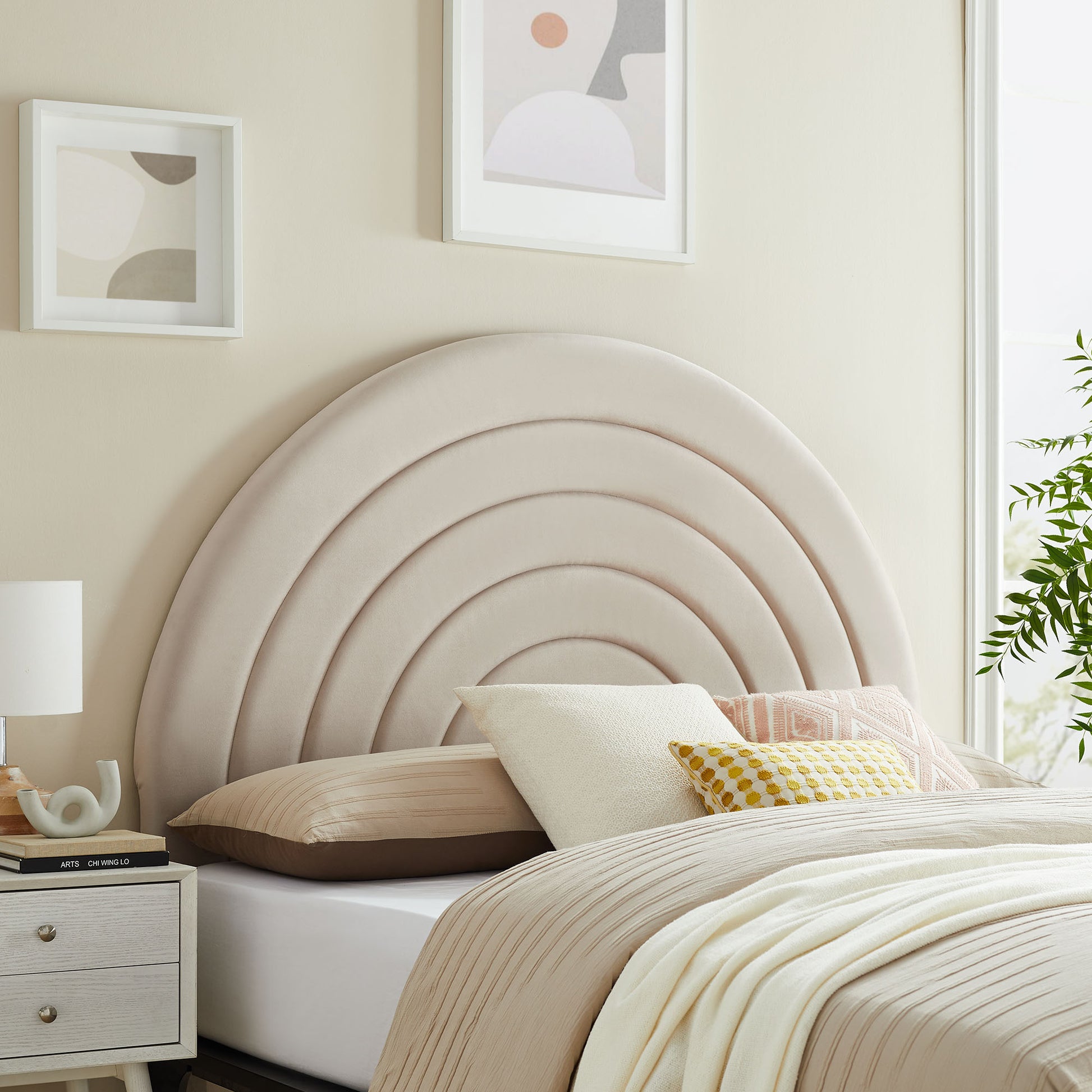 Beige upholstered arch headboard with concentric curves, cream bedding, assorted pillows, bedside lamp, abstract framed art.