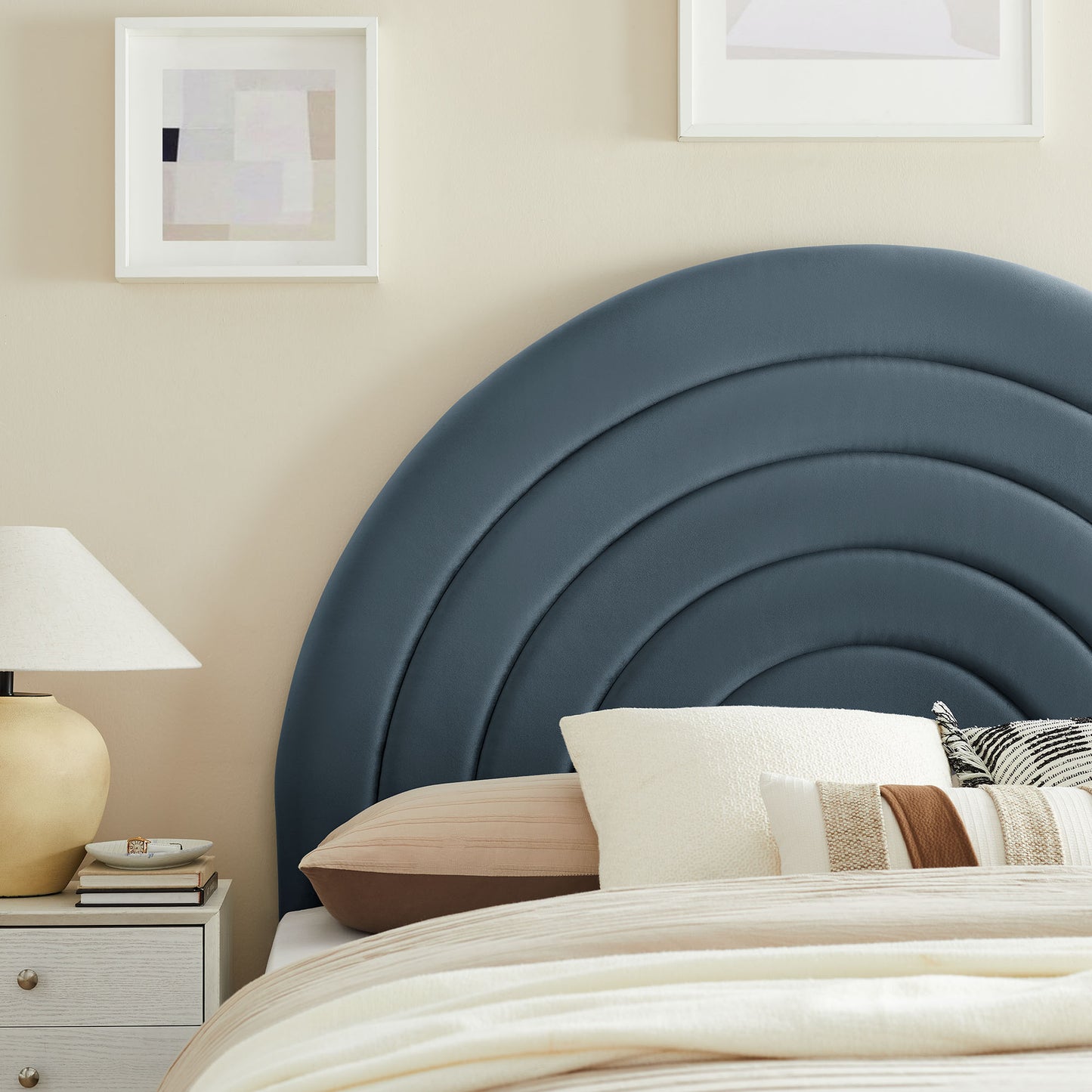 Blue curved upholstered headboard with concentric arches, beige walls, white bedding, striped pillows, lamp, framed art.