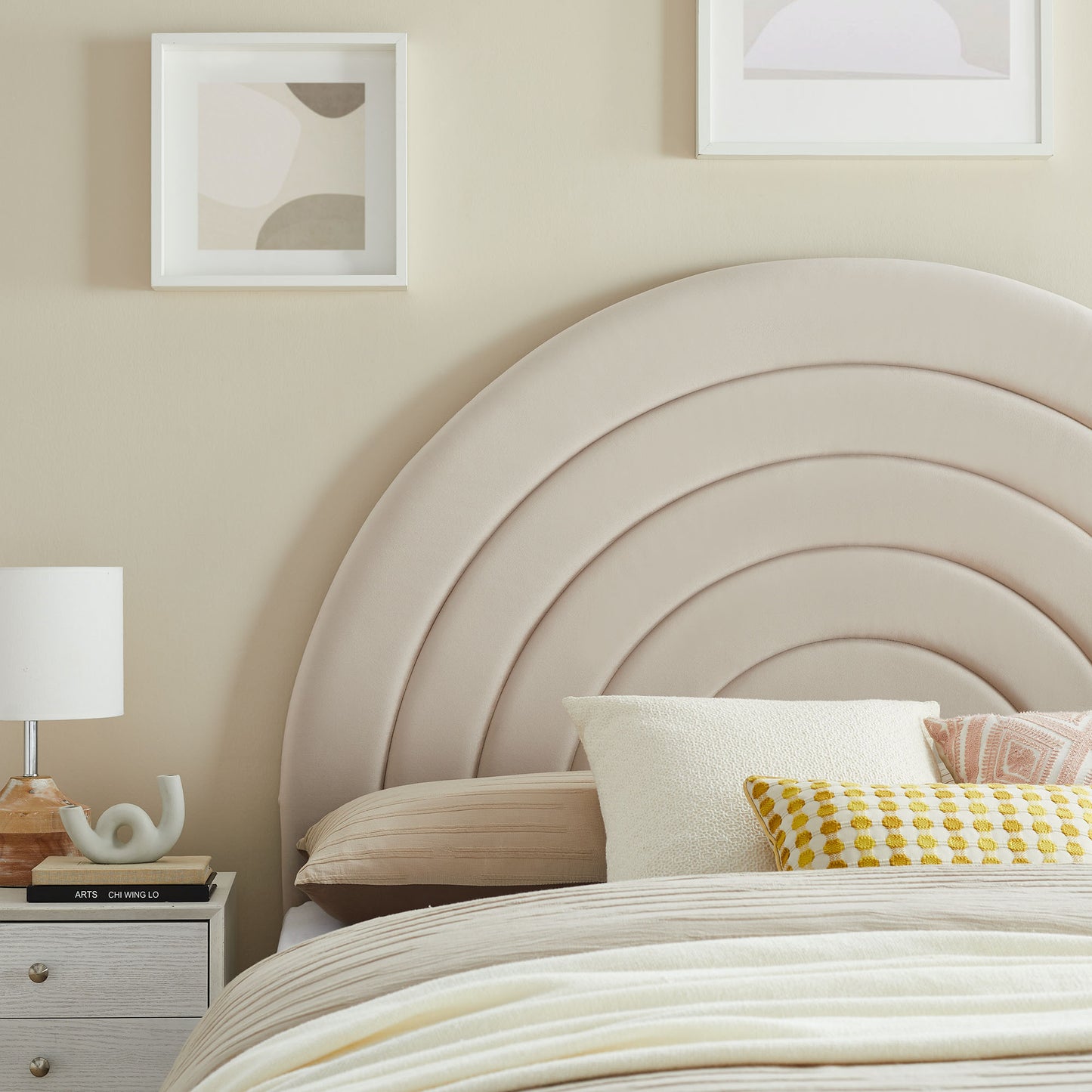 Gray arched velvet headboard with a concentric arc design; beige bedding with white and yellow pillows, and a nightstand with a lamp.