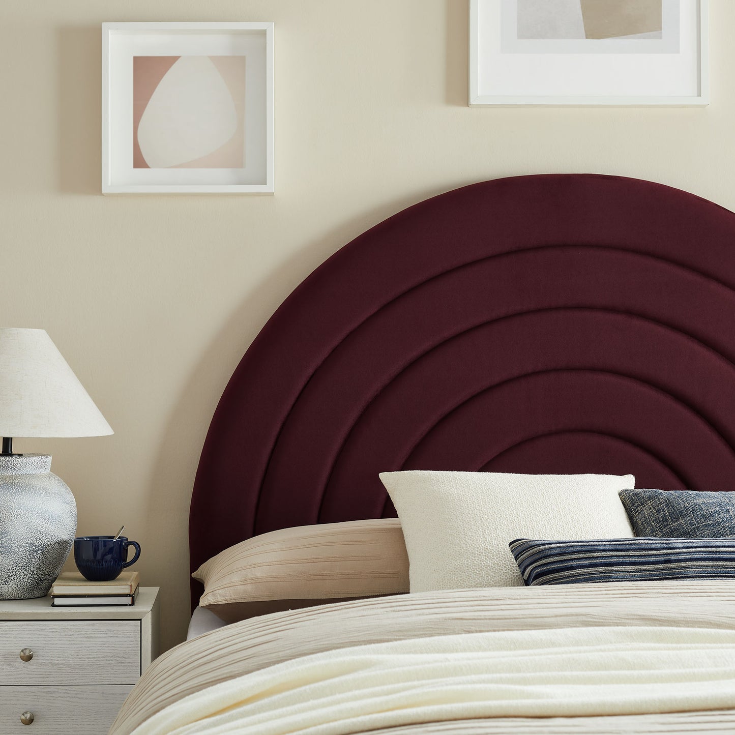 Maroon curved upholstered headboard with concentric arches, beige bedding, white and striped pillows, nightstand lamp.
