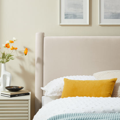 Ivory velvet twin headboard behind a bed with a white quilted duvet & a mustard yellow throw pillow; vase of orange flowers & framed art hung above.
