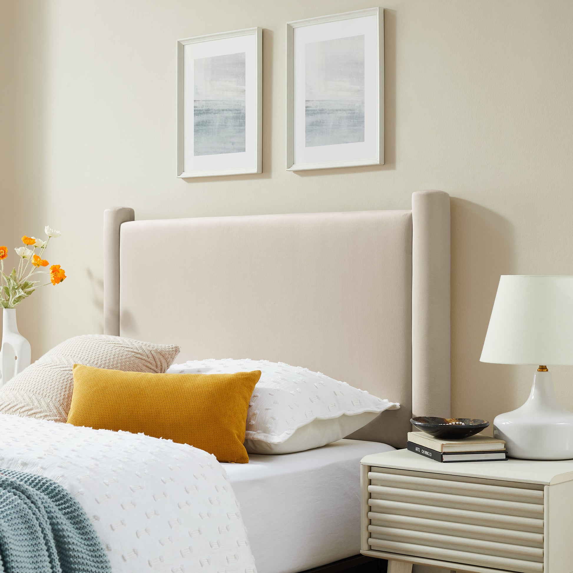 Beige upholstered bed with white duvet, mustard pillow, two framed abstract prints above; white nightstand with lamp.