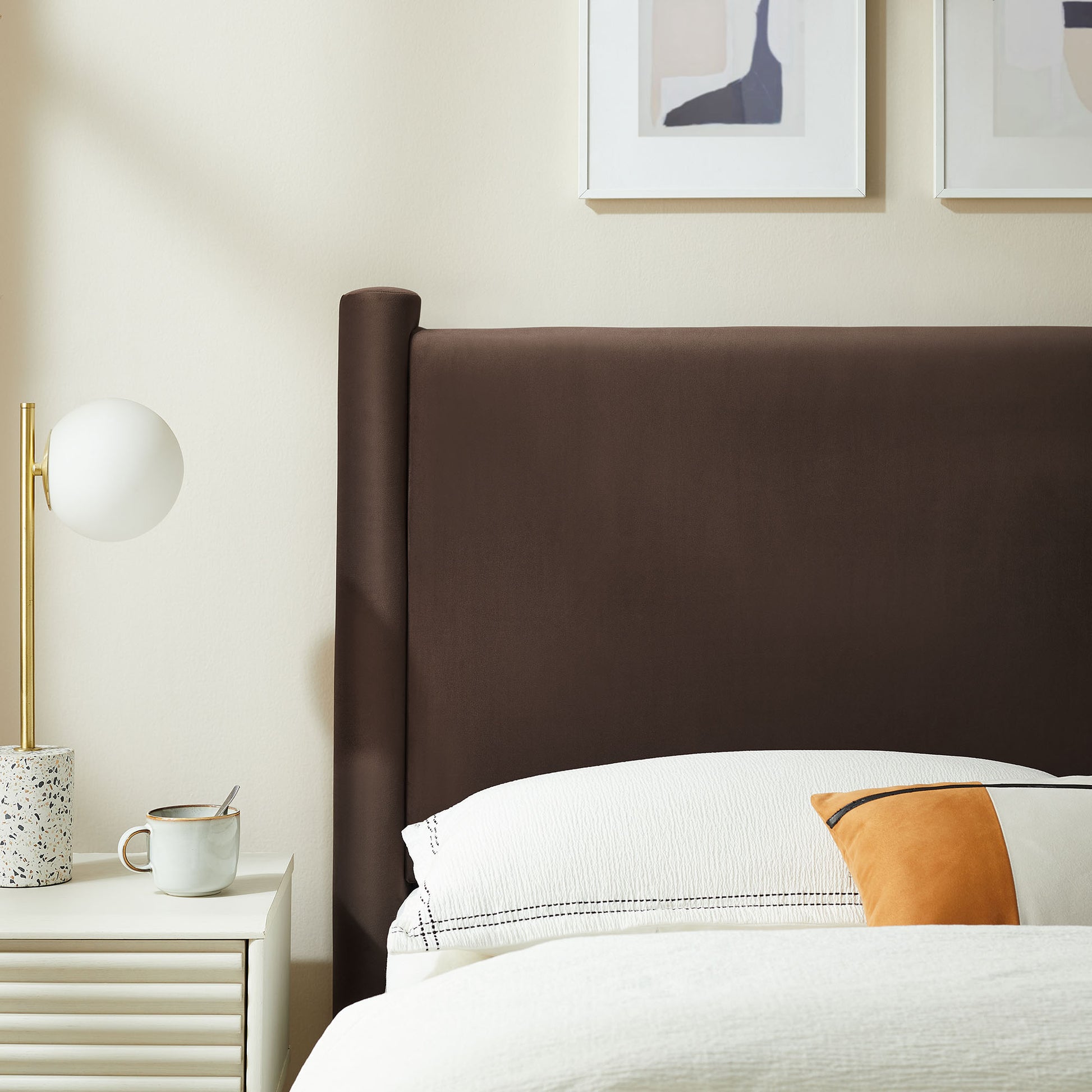 Brown upholstered headboard, white bedding with orange accent pillow, white nightstand, brass globe lamp, abstract art.