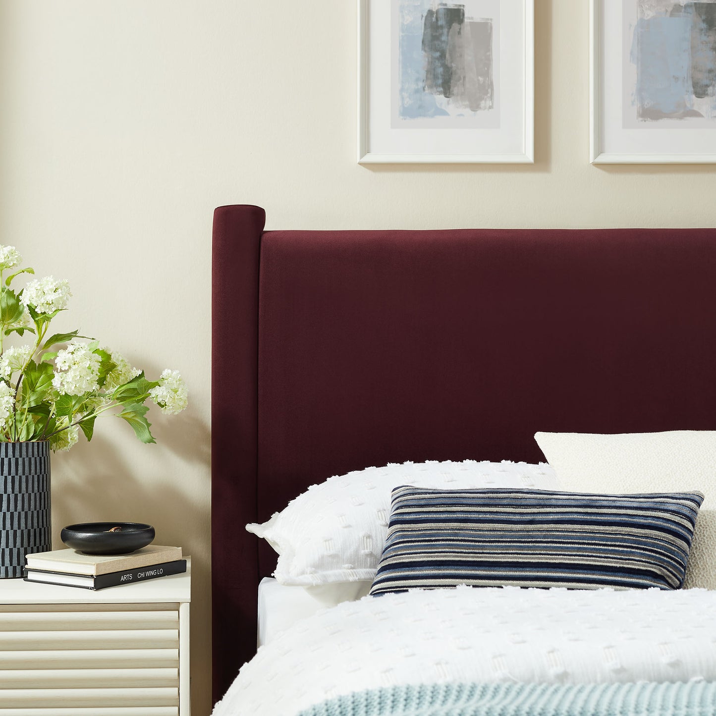 Tall purple velvet headboard with white textured duvet and blue striped pillow; bedside table with flowers.