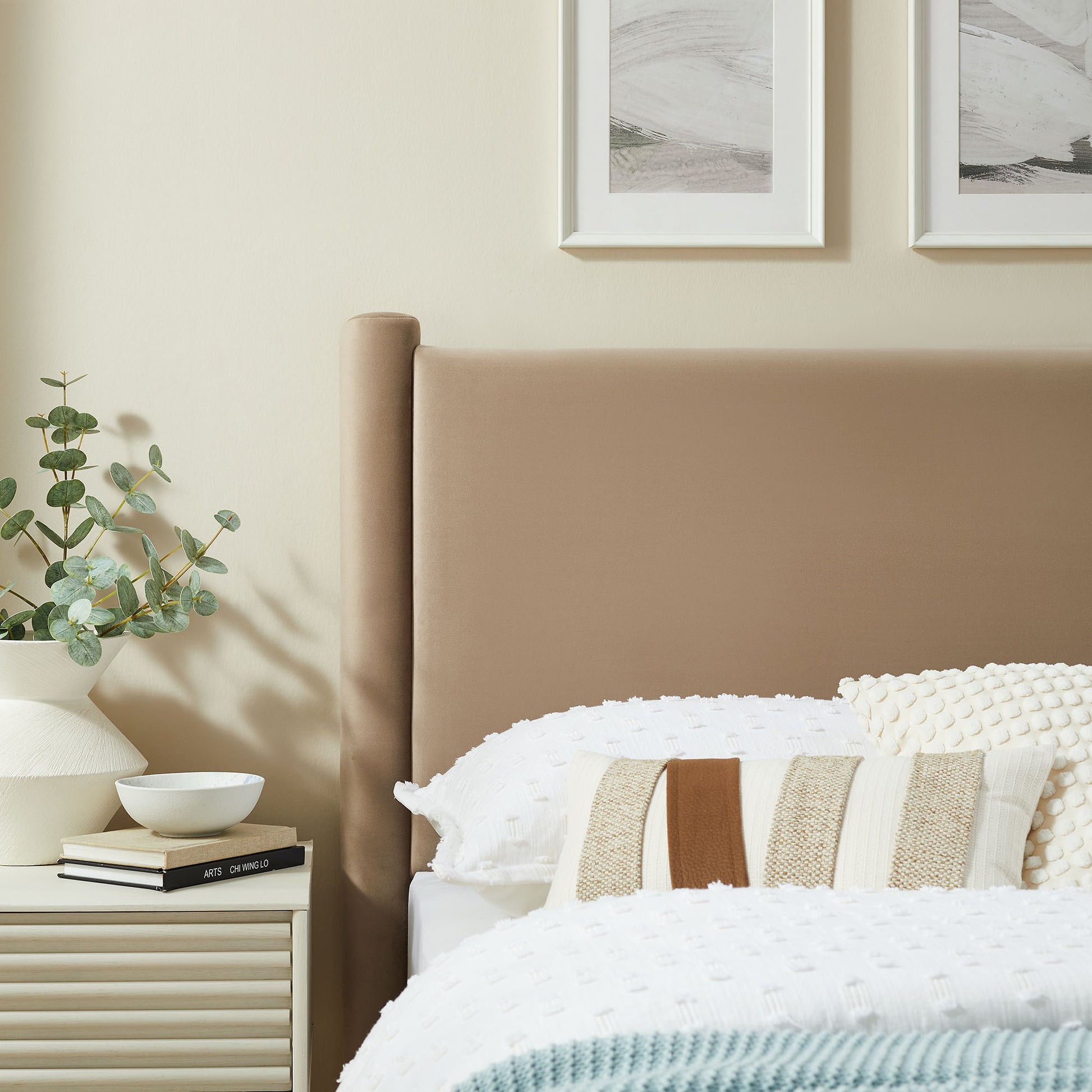 Taupe upholstered twin headboard with white knit bedding, striped pillows, and a cream nightstand holding a plant in a white vase.