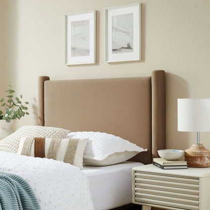 Oatmeal velvet twin headboard with a tall upholstered silhouette; white bedding with textured pillows, prints on the wall, and a beige nightstand with lamp.