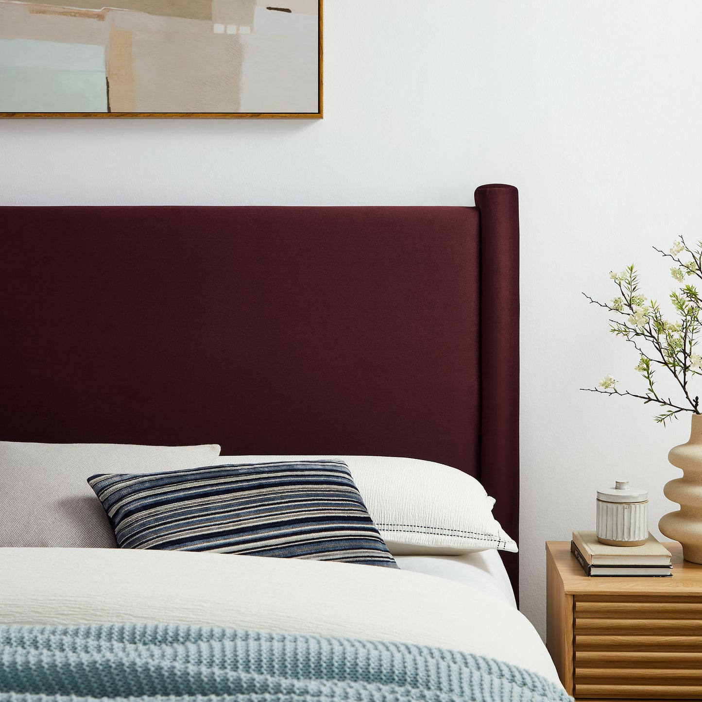 Purple upholstered full headboard with white bedding, blue striped pillow, light blue throw; wood nightstand with plant.