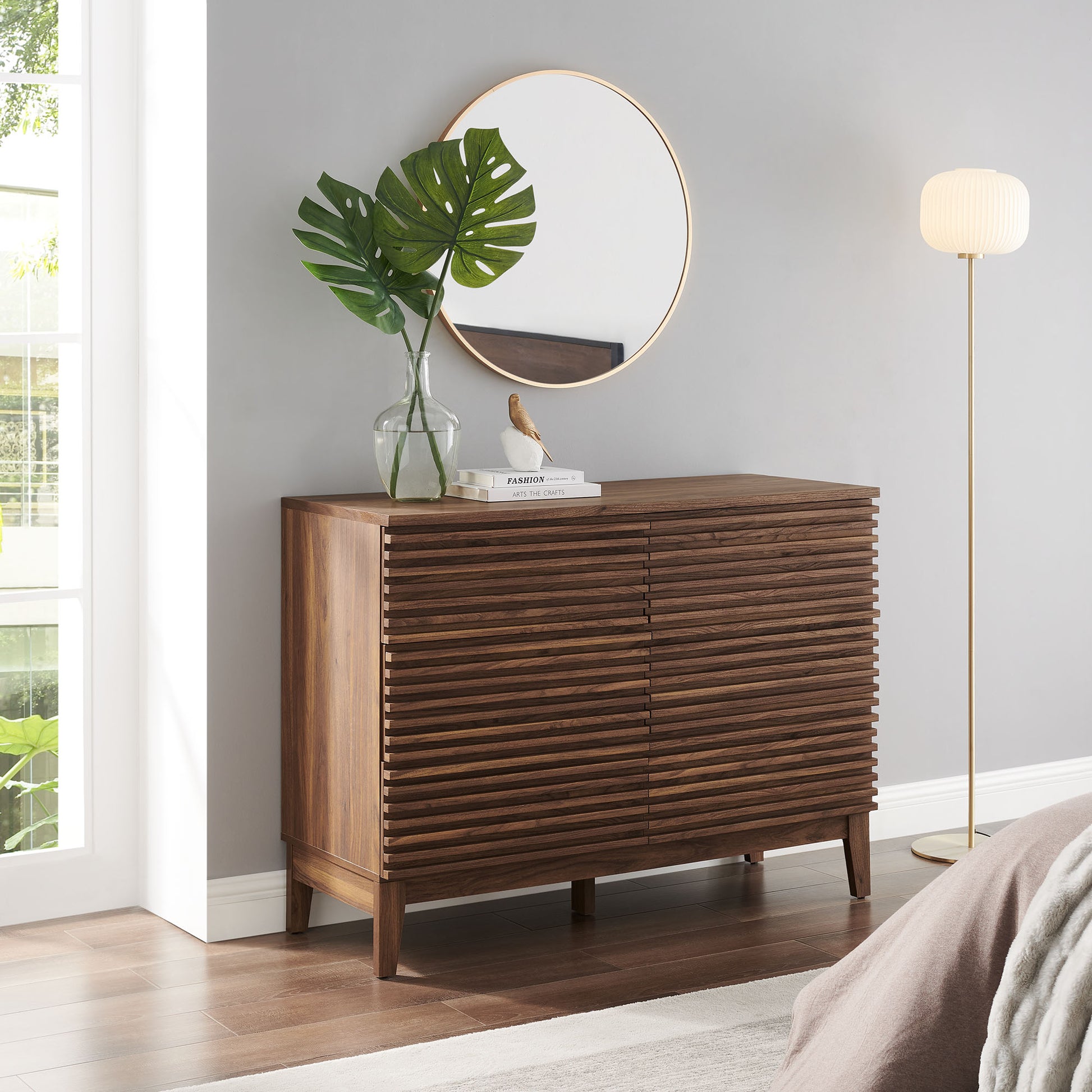 Walnut slatted 6-drawer dresser with circular gold-rim mirror, glass vase with monstera leaf, books; floor lamp nearby.