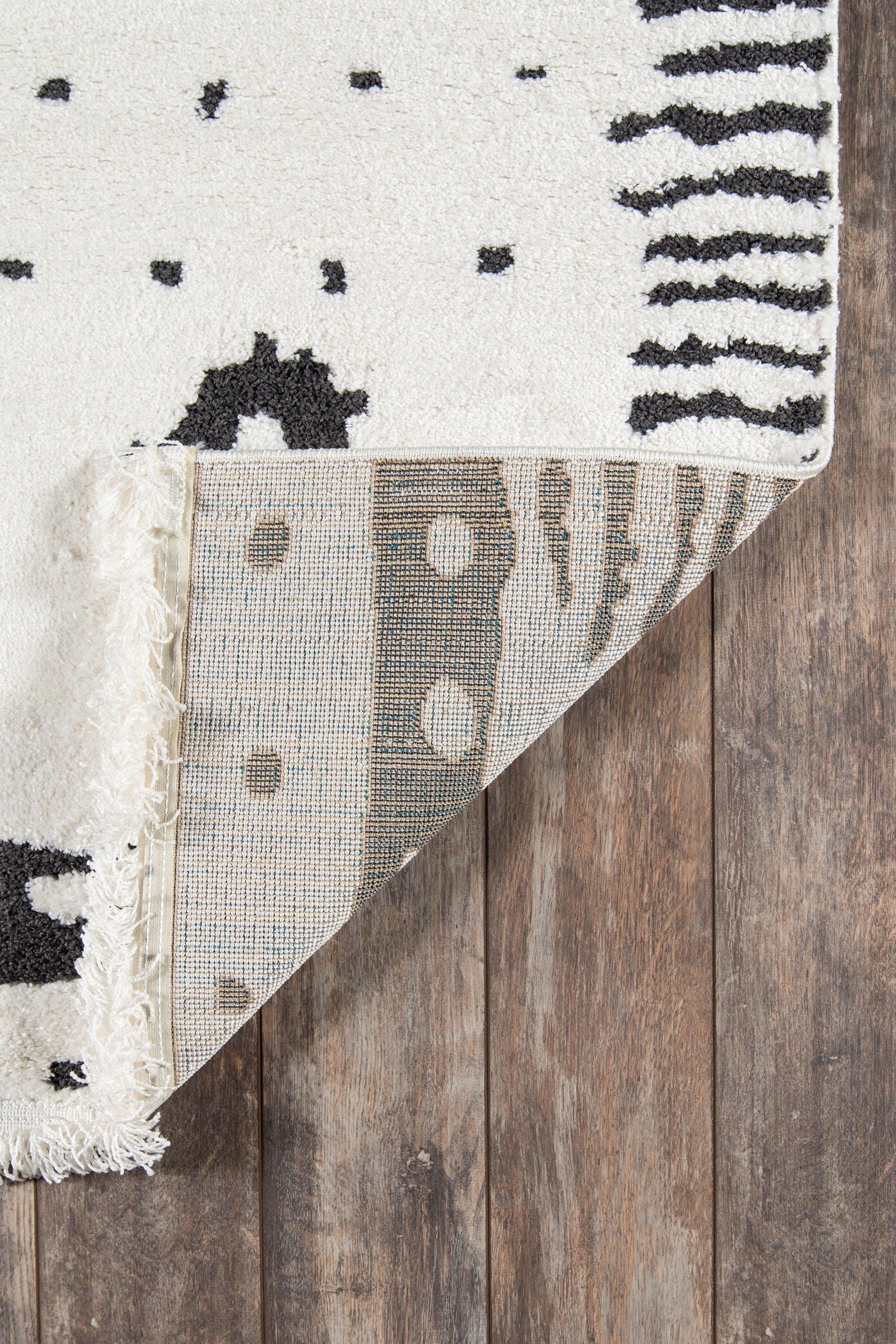 Ivory cotton area rug with black geometric dots and stripes, fringed edge, shown on a wood floor.