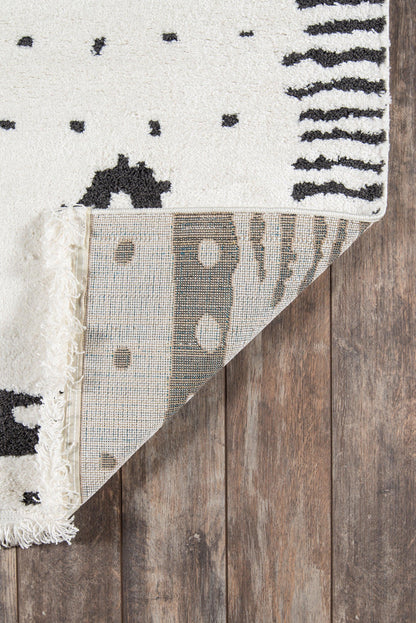 Ivory cotton area rug with black geometric dots and stripes, fringed edge, shown on a wood floor.
