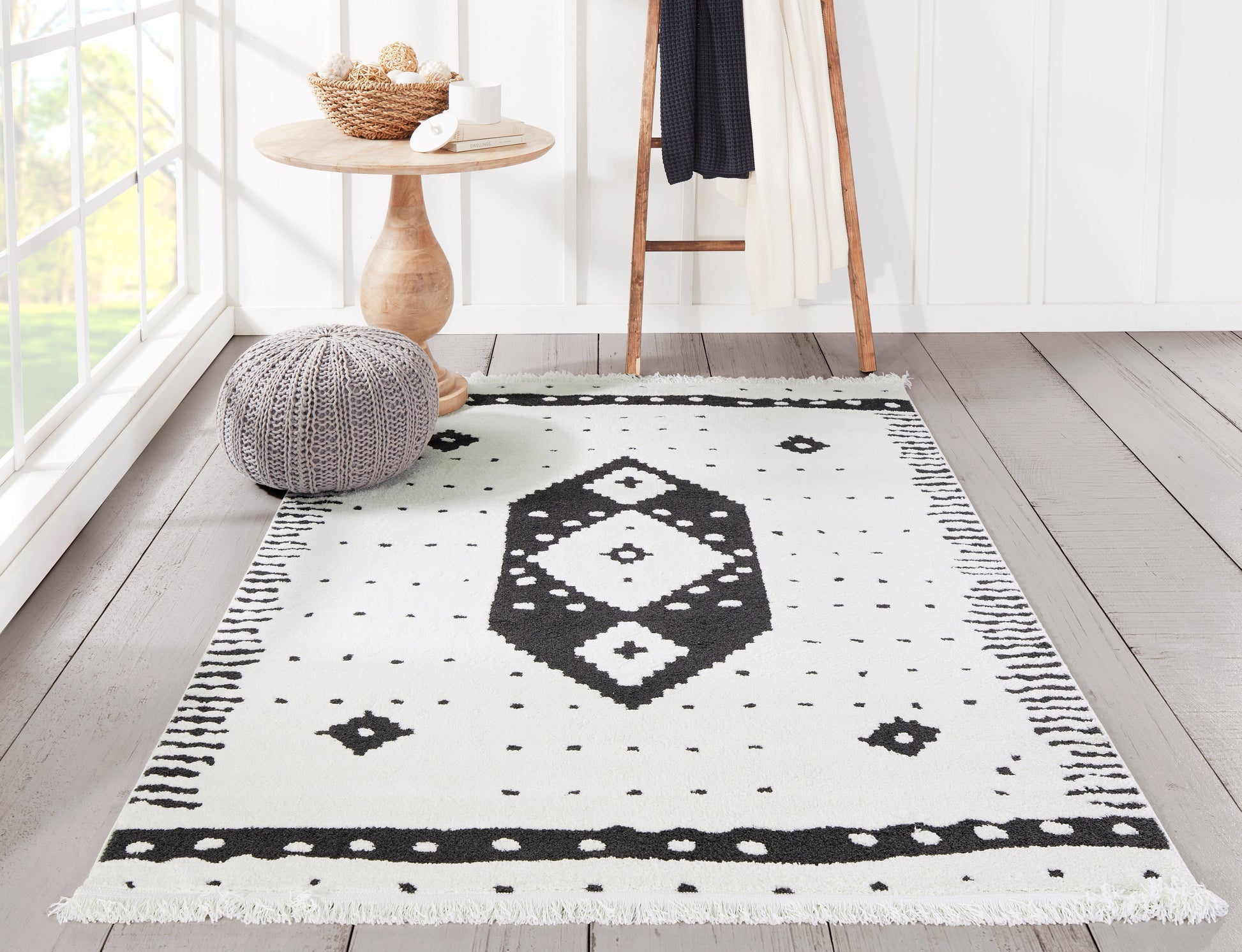 White rug with black geometric diamond motif and fringed edges in a bright room with a wooden table and knit pouf.