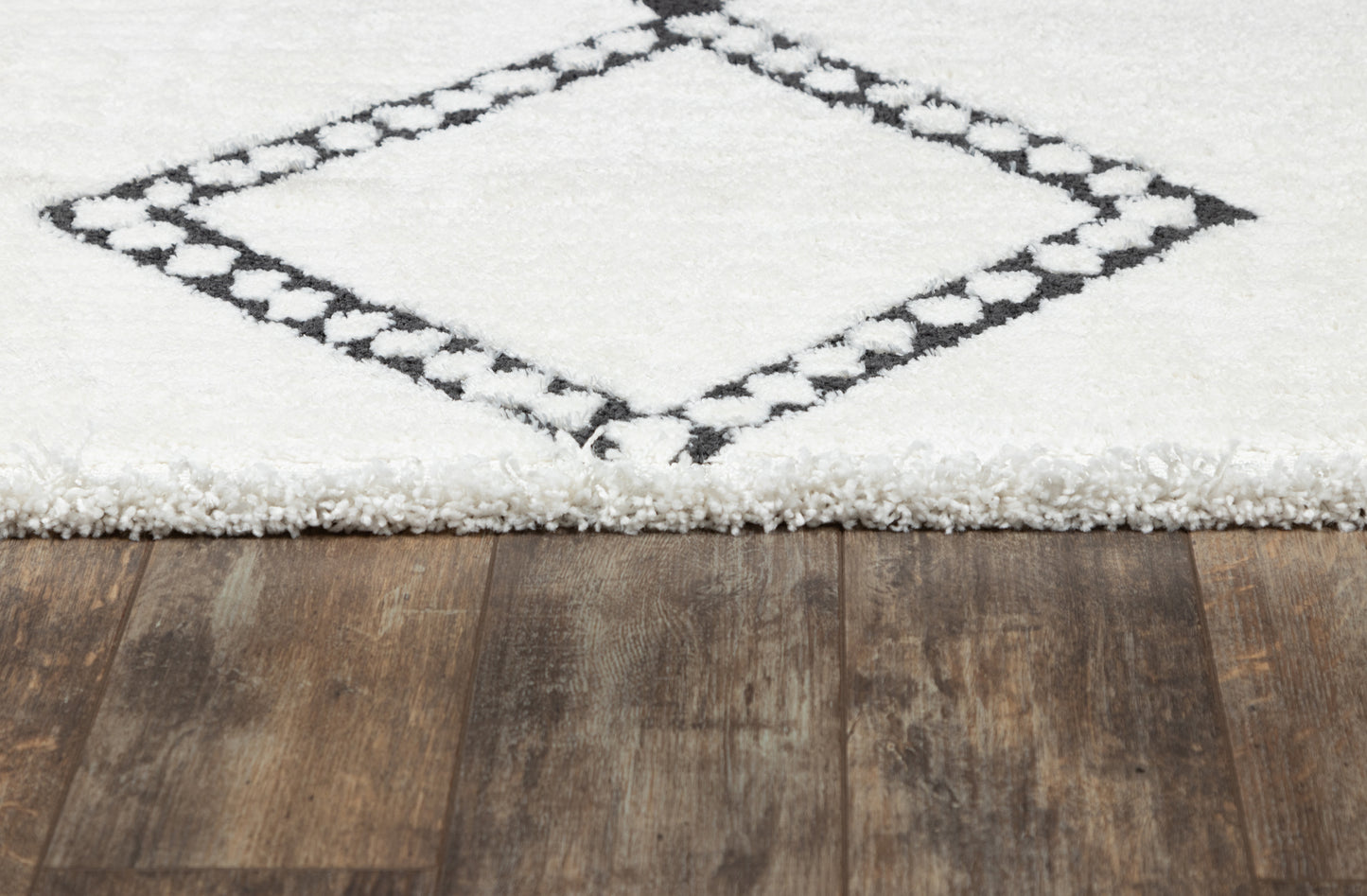 White shag rug with a black diamond border on a wooden floor, fringed edge.