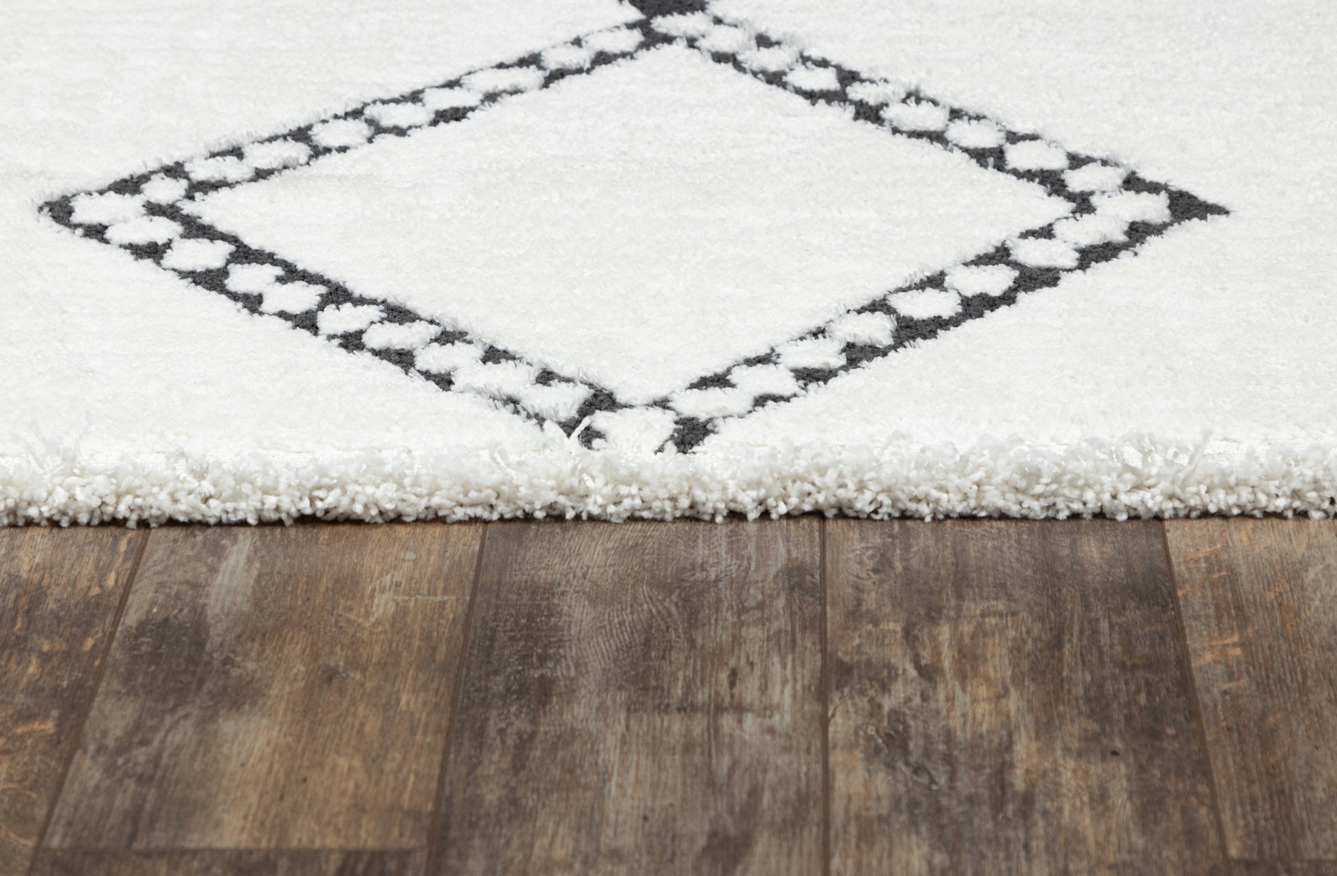 White shag rug with a black diamond border on a wooden floor, fringed edge.