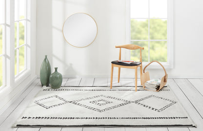 Minimalist living room rug with black diamond border, light wood chair, gold-framed mirror, green vases, wicker basket.