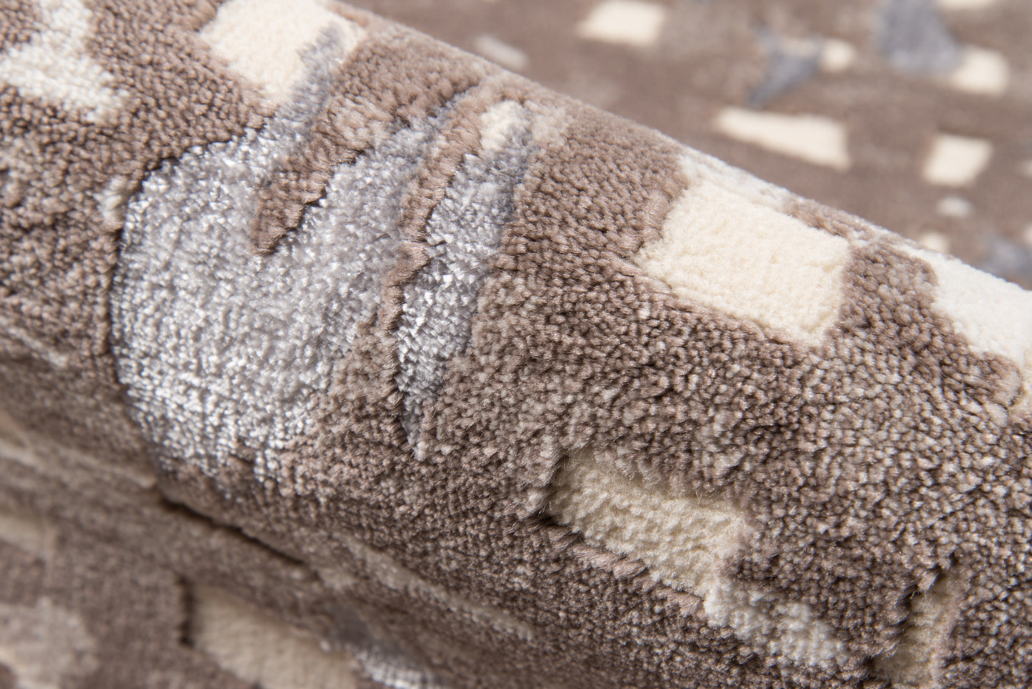 Close-up of a plush shag rug in brown, beige & cream with a geometric square pattern; rolled edge shows soft, dense pile.