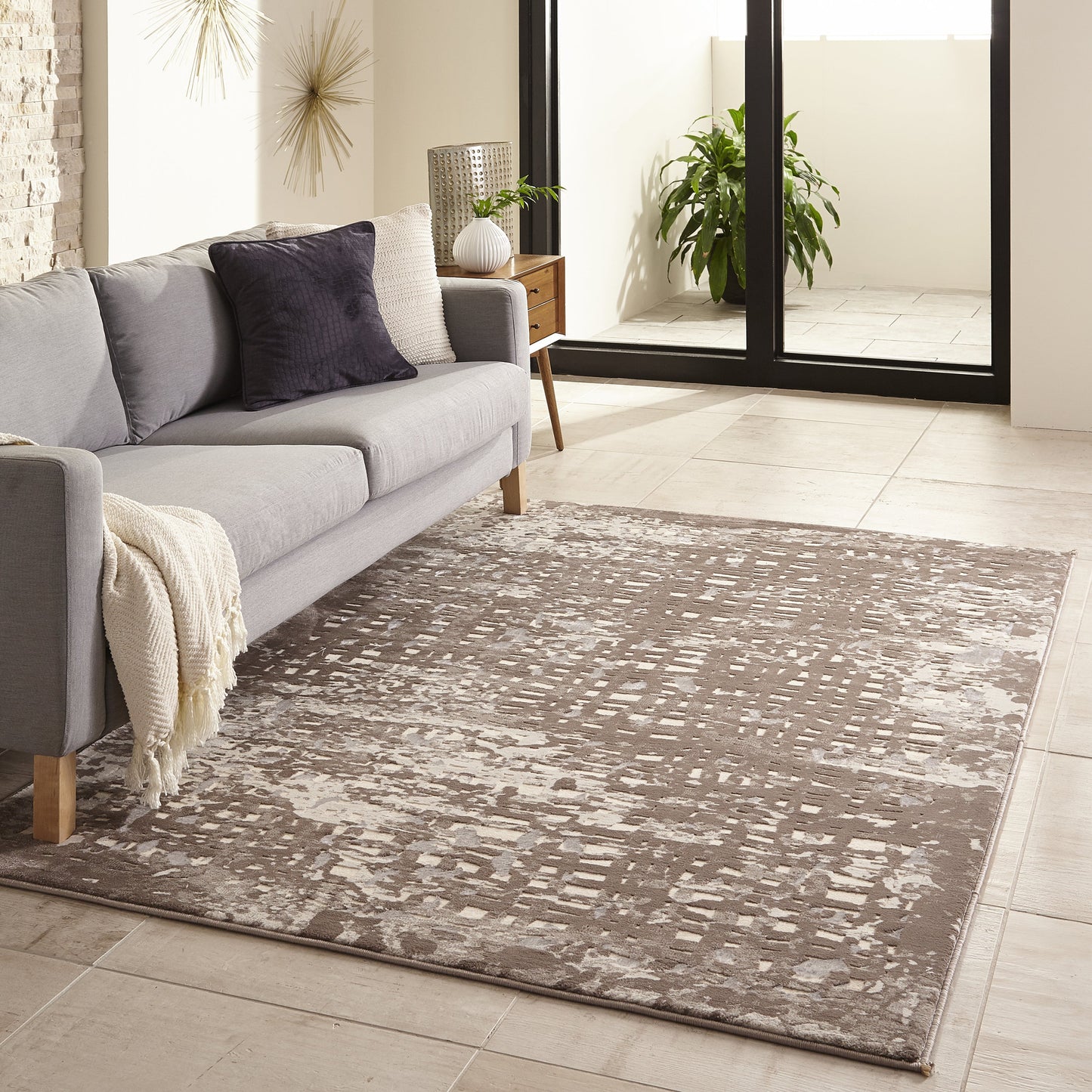 Gray fabric sofa with wooden legs on a taupe abstract rug; beige throw, dark pillow, sunlit living room with glass doors.