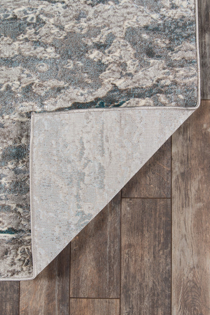 Teal, gray and beige patterned area rug with distressed design, folded corner shows beige underside; hardwood floor backdrop.