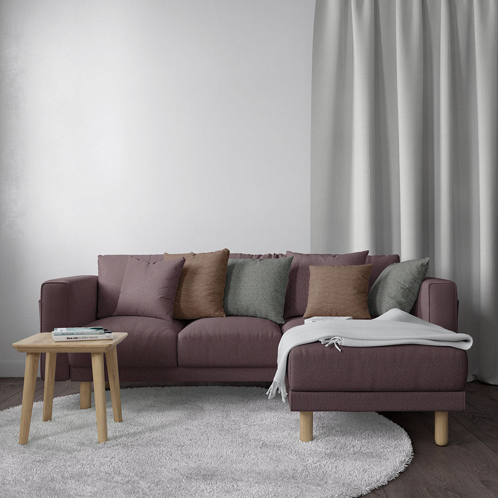 Dusty mauve L-shaped fabric sofa with chaise, earth-toned cushions, light wood legs, gray throw, beside a round coffee table.