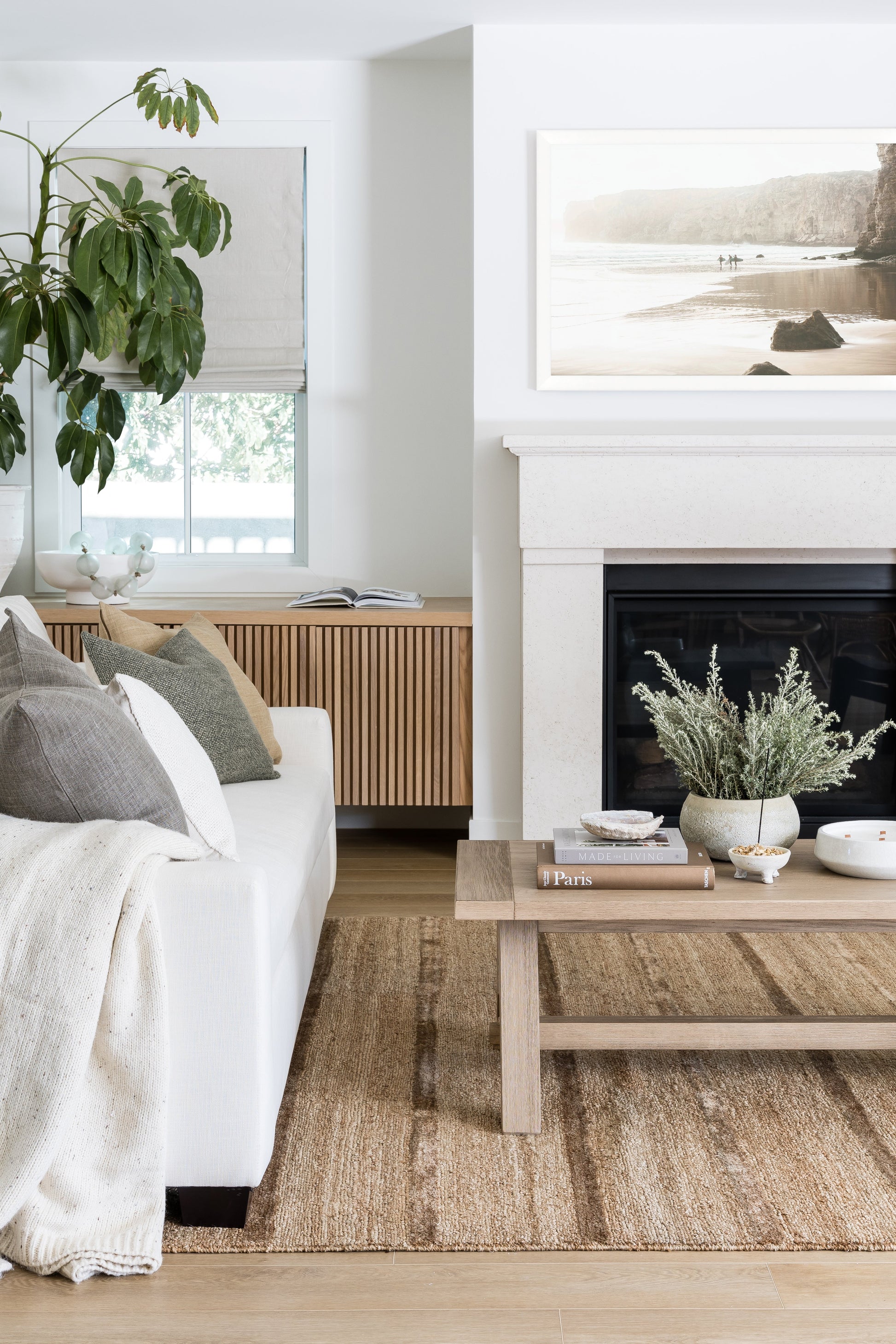 Bright living room with white sofa, beige throw, cushions, natural wood coffee table, potted plant, jute rug, fireplace.