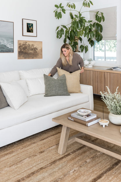 Bright living room with white sofa, beige and gray cushions, a wooden coffee table, large potted plant, wall art, and a rug.