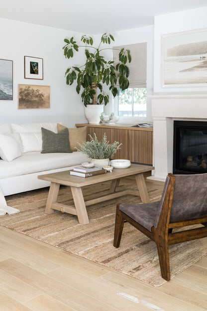 Natural wood coffee table in a light, airy living room with a large potted plant, white sofa, beige rug, and fireplace.