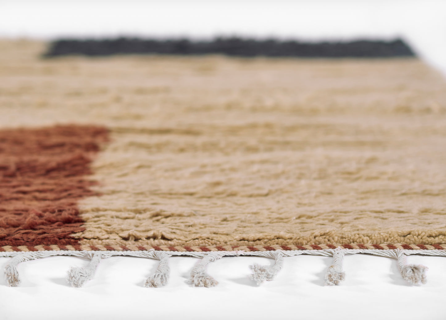 Close-up of a woven rug with beige field, rust red stripe, dark blue border, and white tassel fringe along the edge.