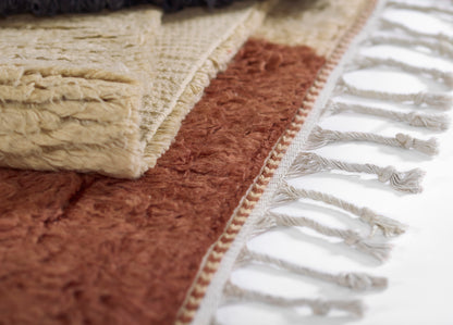 Beige plush area rug with tassel fringe over a rust shag rug; layered look with cream tassels along the edge.