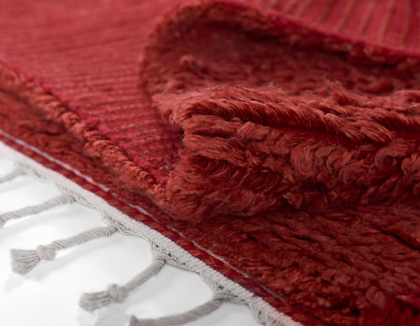Red shag rug with plush pile and rolled edge, tassel fringe along a white surface.