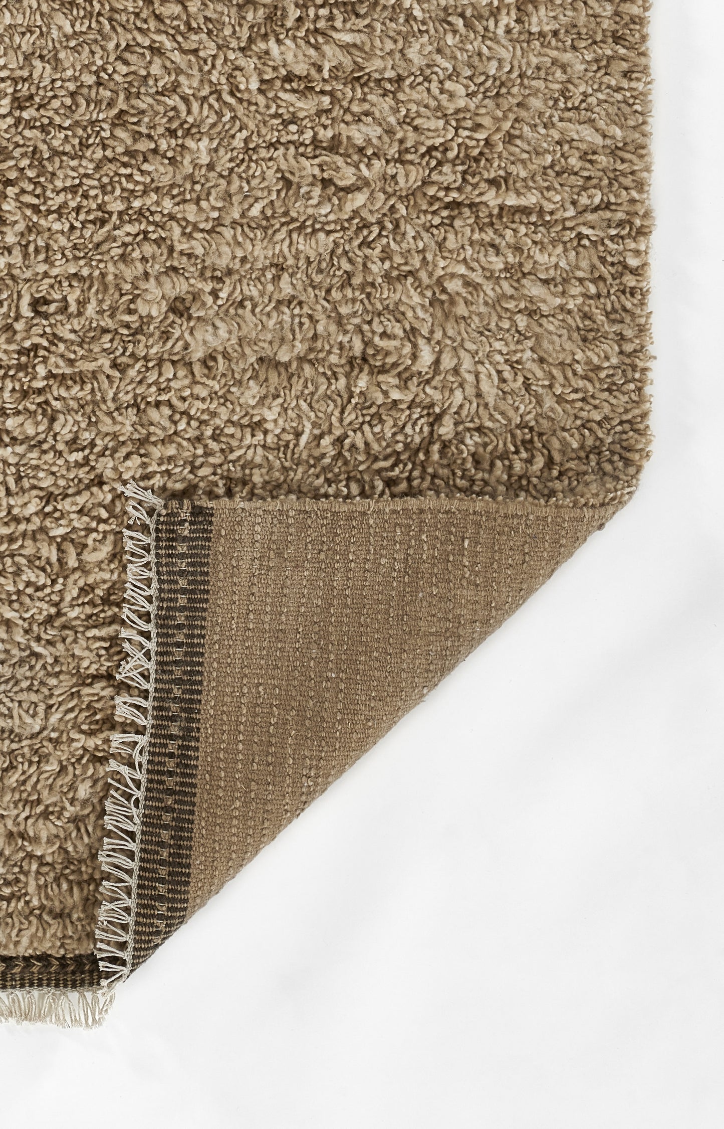 Beige-brown shag rug with fluffy pile; corner folded to expose brown woven backing and white fringe trim.