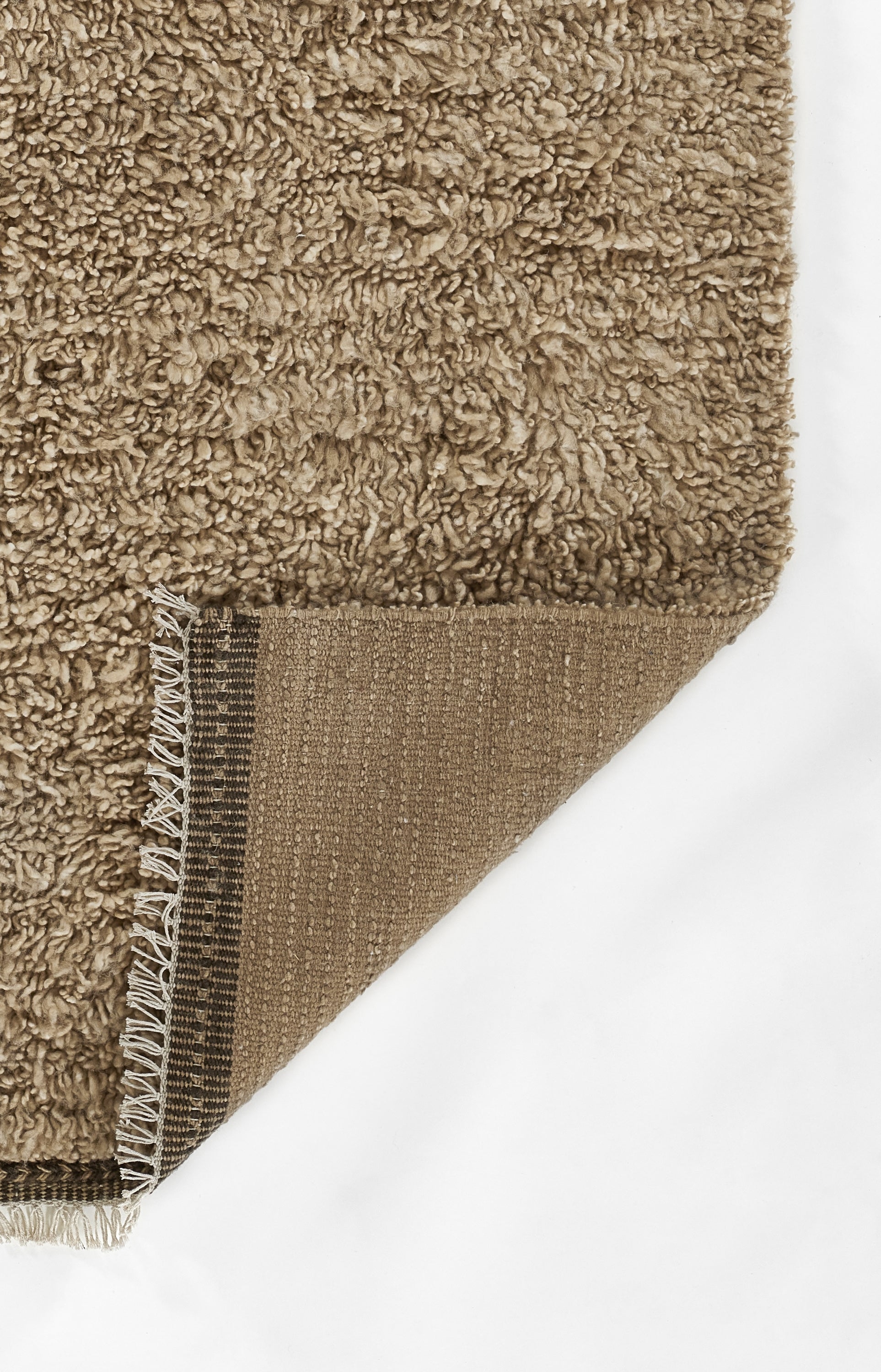 Beige-brown shag rug with fluffy pile; corner folded to expose brown woven backing and white fringe trim.