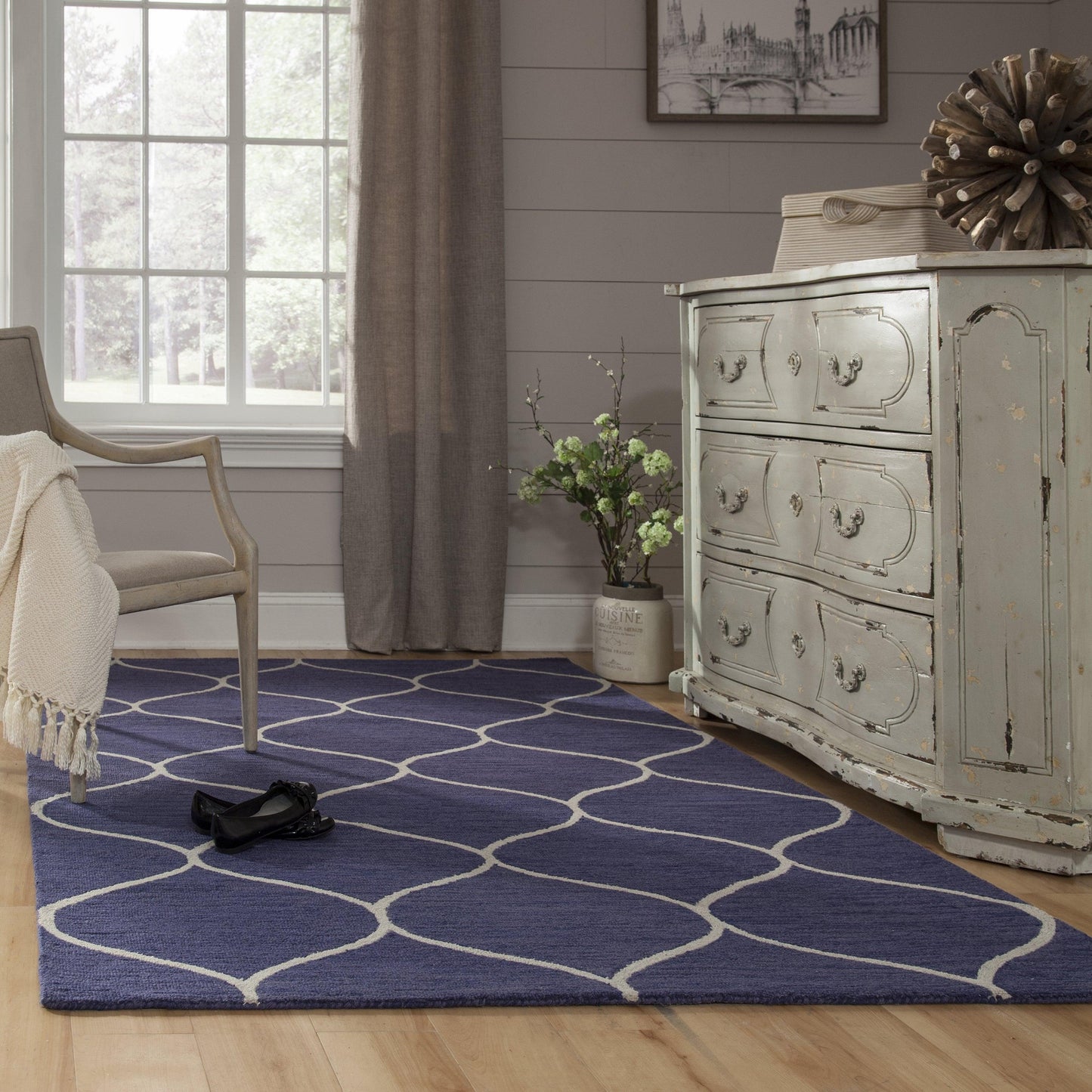 Blue trellis-pattern area rug on hardwood floor in a cozy living room with distressed gray dresser & beige armchair.
