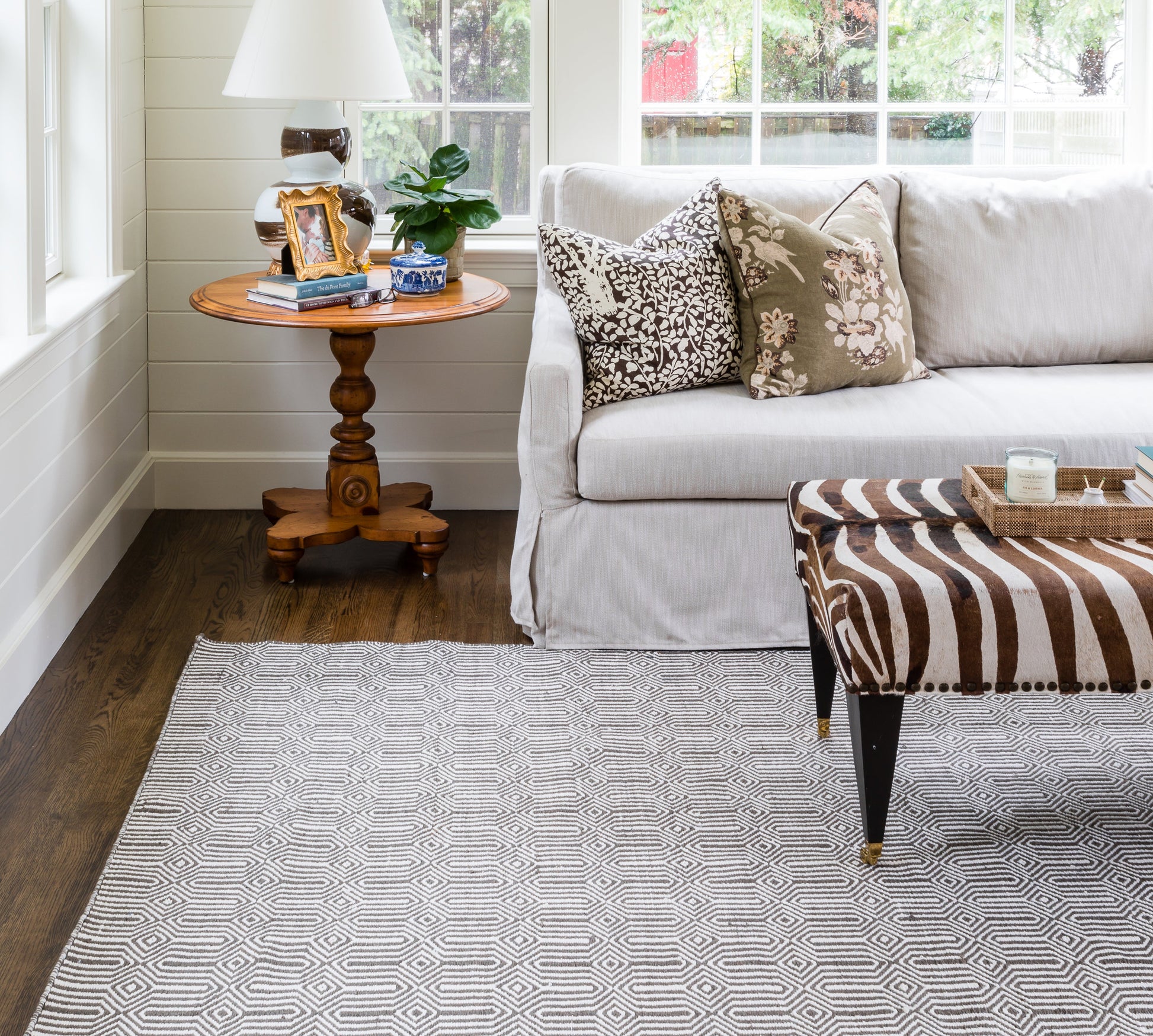 Cozy living room: light gray sofa, botanical cushions, wooden pedestal table with lamp, plant; zebra ottoman; geometric rug.