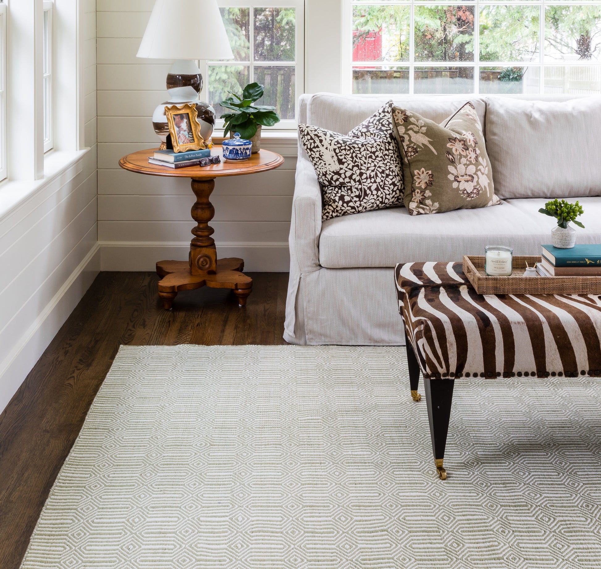 Cozy living room with light gray sofa, patterned pillows, round wood pedestal table, lamp, plant, zebra-print ottoman, rug.