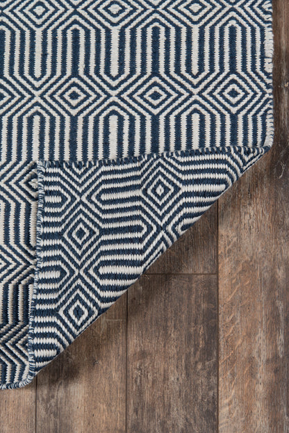 Navy and white geometric rug with diamond motifs and fringed edge, folded corner reveals underside on wood floor.