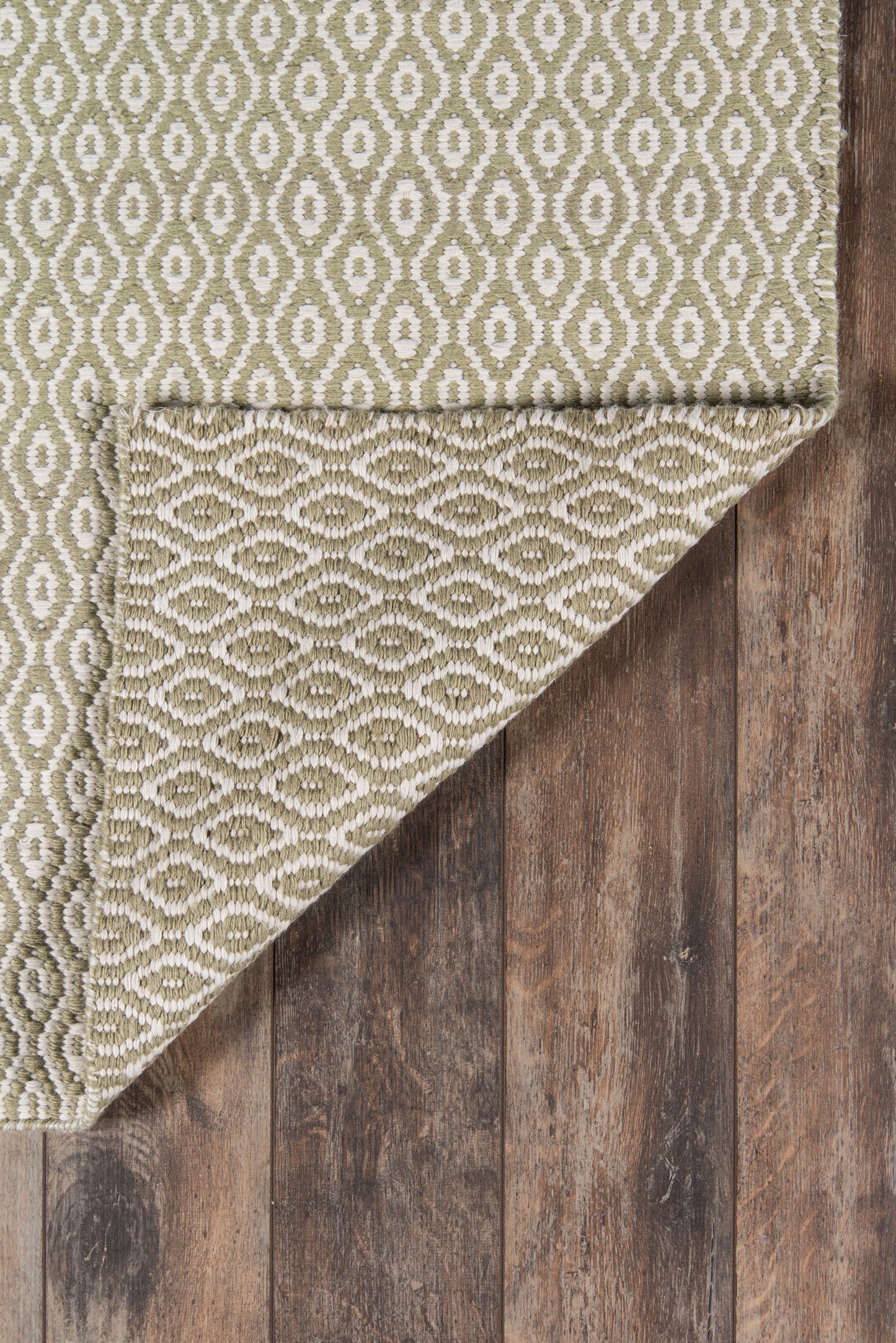 Olive-green geometric rug with ivory diamond motifs, woven texture, folded corner on rustic wood floor.