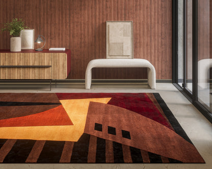 Geometric area rug in orange, yellow and brown tones with a white curved bench beside a wood-paneled wall and glass doors.