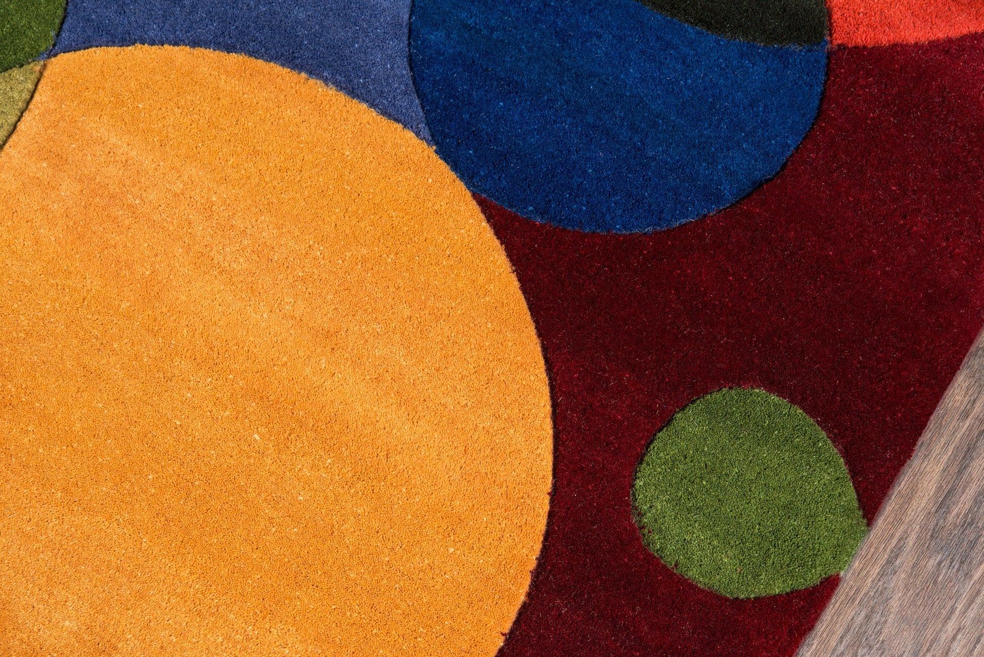 Close-up of a colorful abstract rug: orange, blue and green circles on burgundy background, plush pile near a wood floor edge.