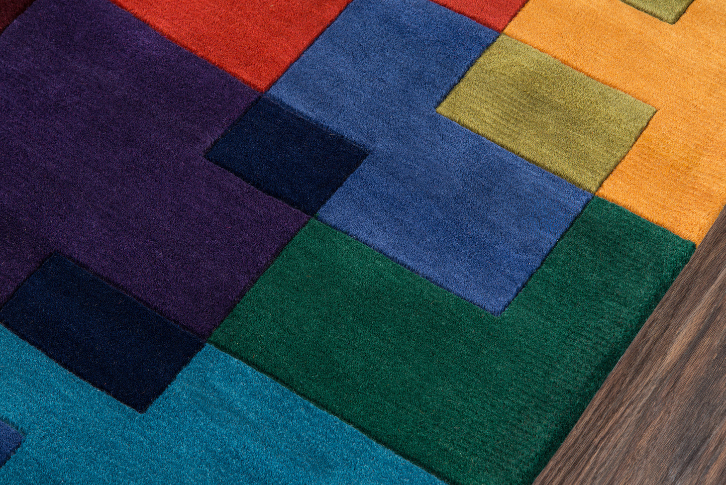 Colorful patchwork rug with square blocks in red, purple, blue, green, yellow and orange; wood floor edge visible.